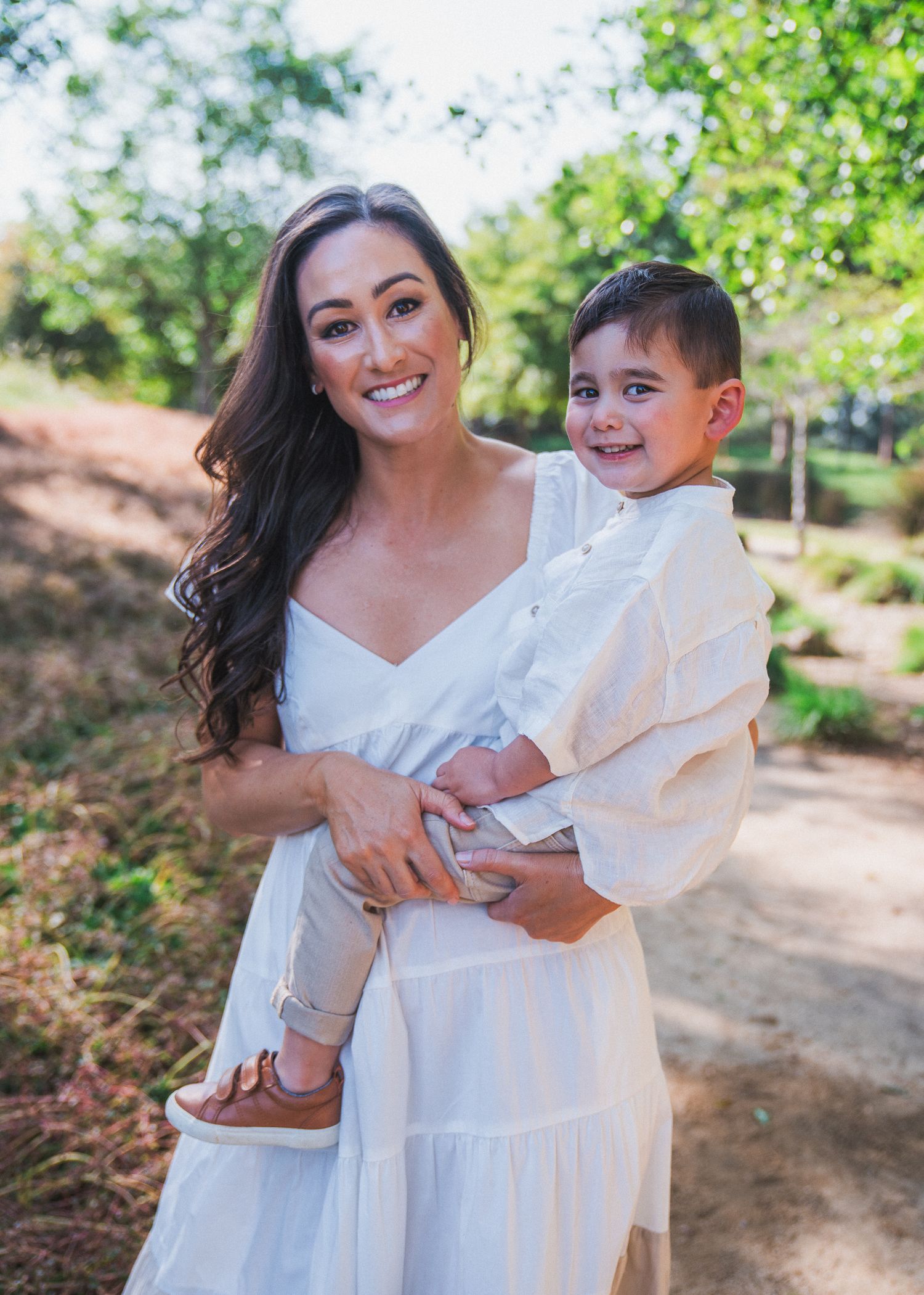 Woman in flowing white dress holds young boy wearing white on garden path surrounded by greenery.
