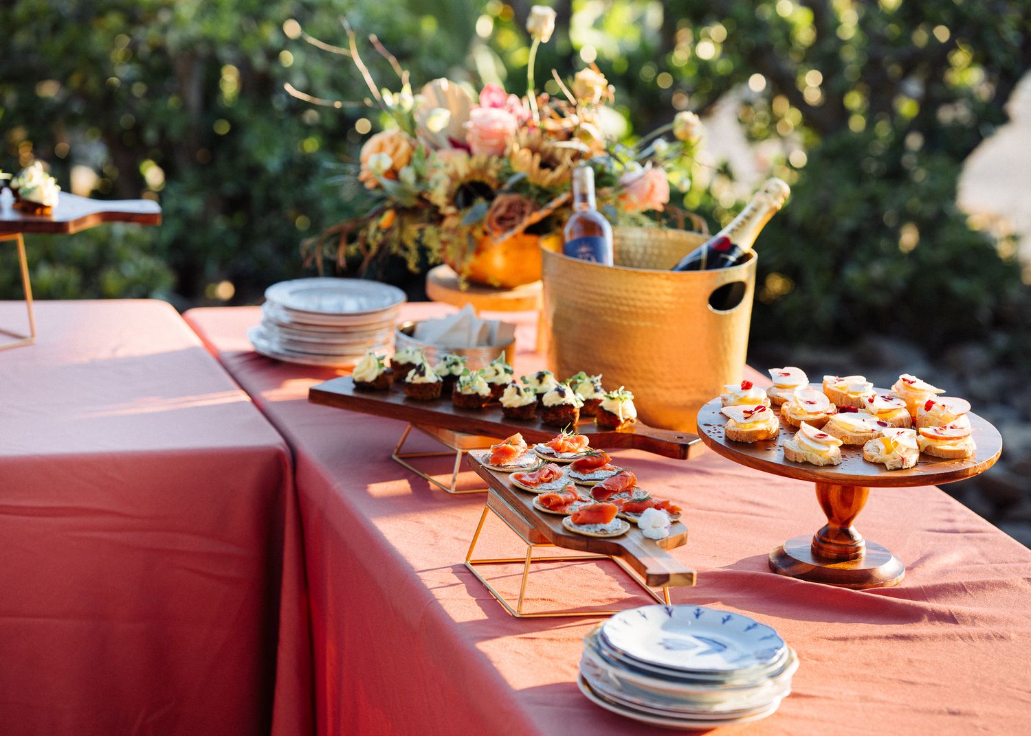 Catering spread on coral tablecloth with cake stands, floral arrangements, and appetizer plates.
