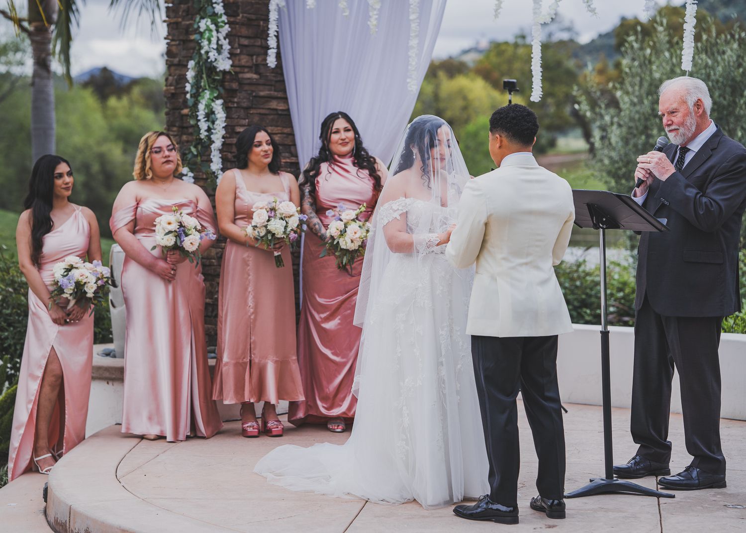 Wedding party poses outdoors in coordinated blush pink and formal attire during elegant daytime ceremony.