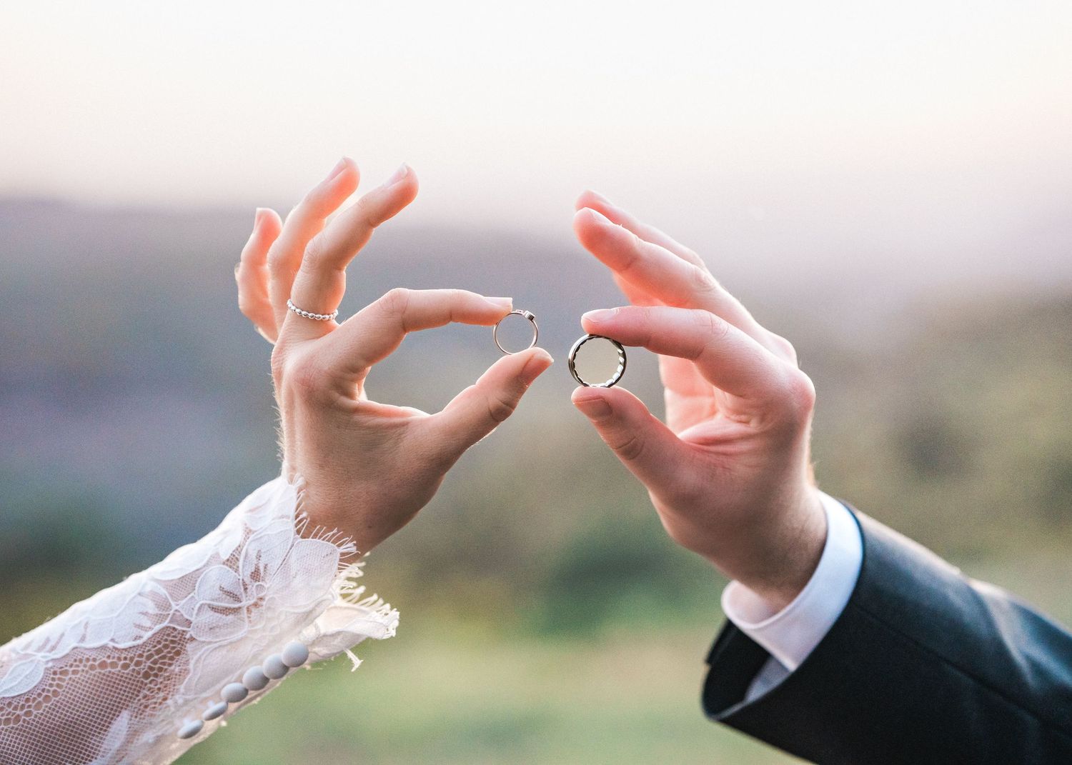 Close-up of hands holding wedding rings against a blurred mountainous background.