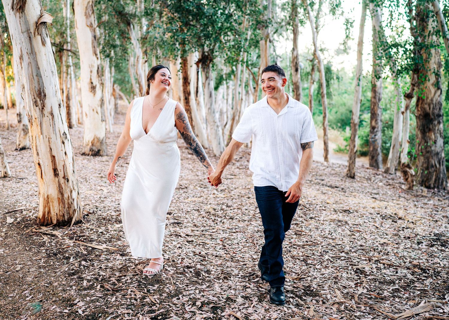 A couple in white and navy outfits walks together through a eucalyptus grove on a sunny day.