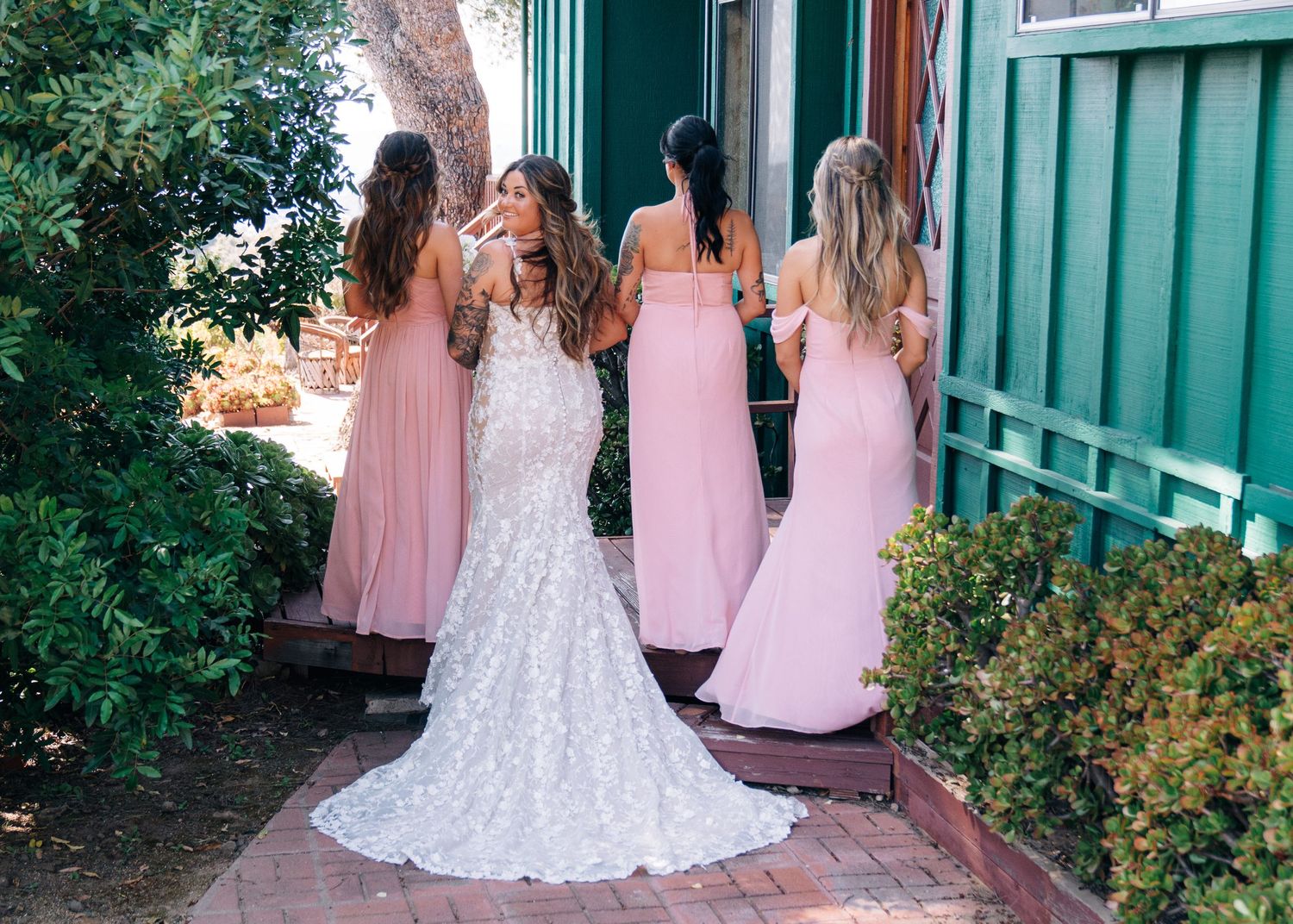 A person in white lace wedding gown surrounded by bridesmaids wearing pink dresses in an outdoor garden setting.