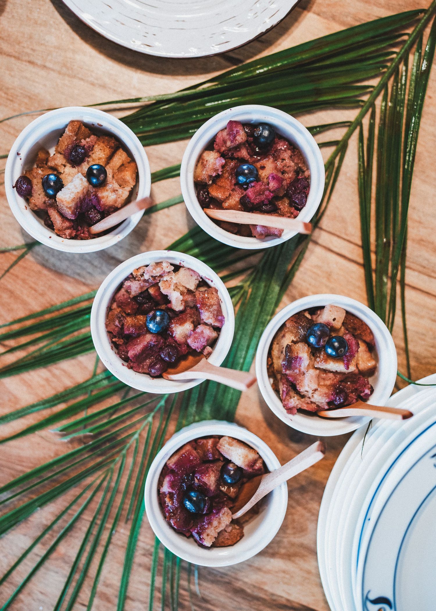 Five white bowls filled with bread pudding topped with fresh blueberries arranged on a wooden table with palm fronds.