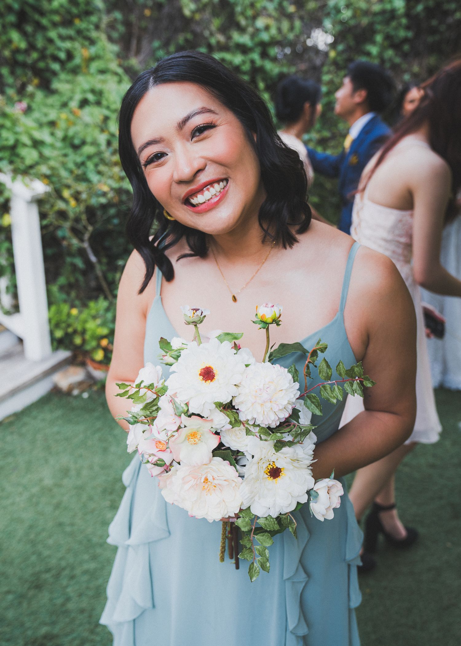 A joyful person in a light blue bridesmaid dress holds a beautiful white and peach floral bouquet at an outdoor wedding.