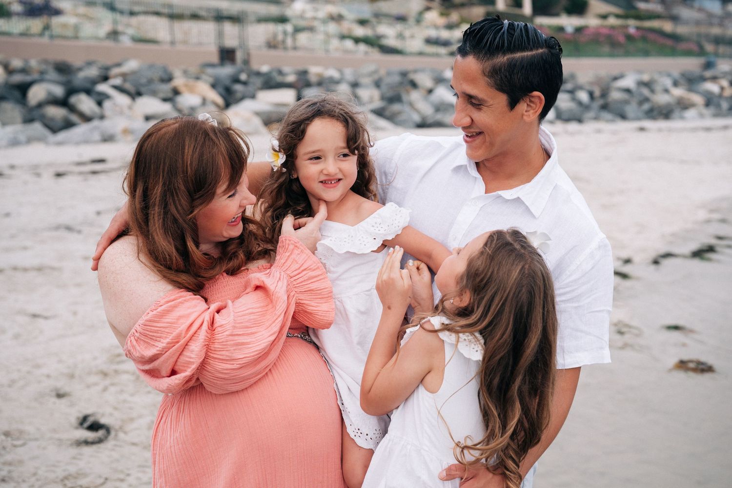 Family in coordinated pink and white outfits enjoying a playful moment together on a scenic beach.