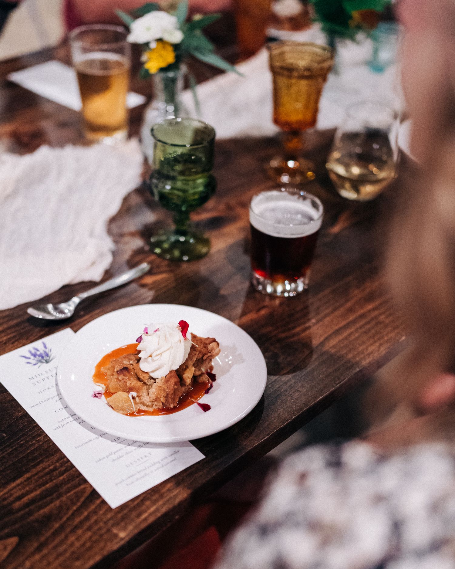 A dessert plate with whipped cream topped pastry sits on a wooden table alongside various colorful drinks in glassware.