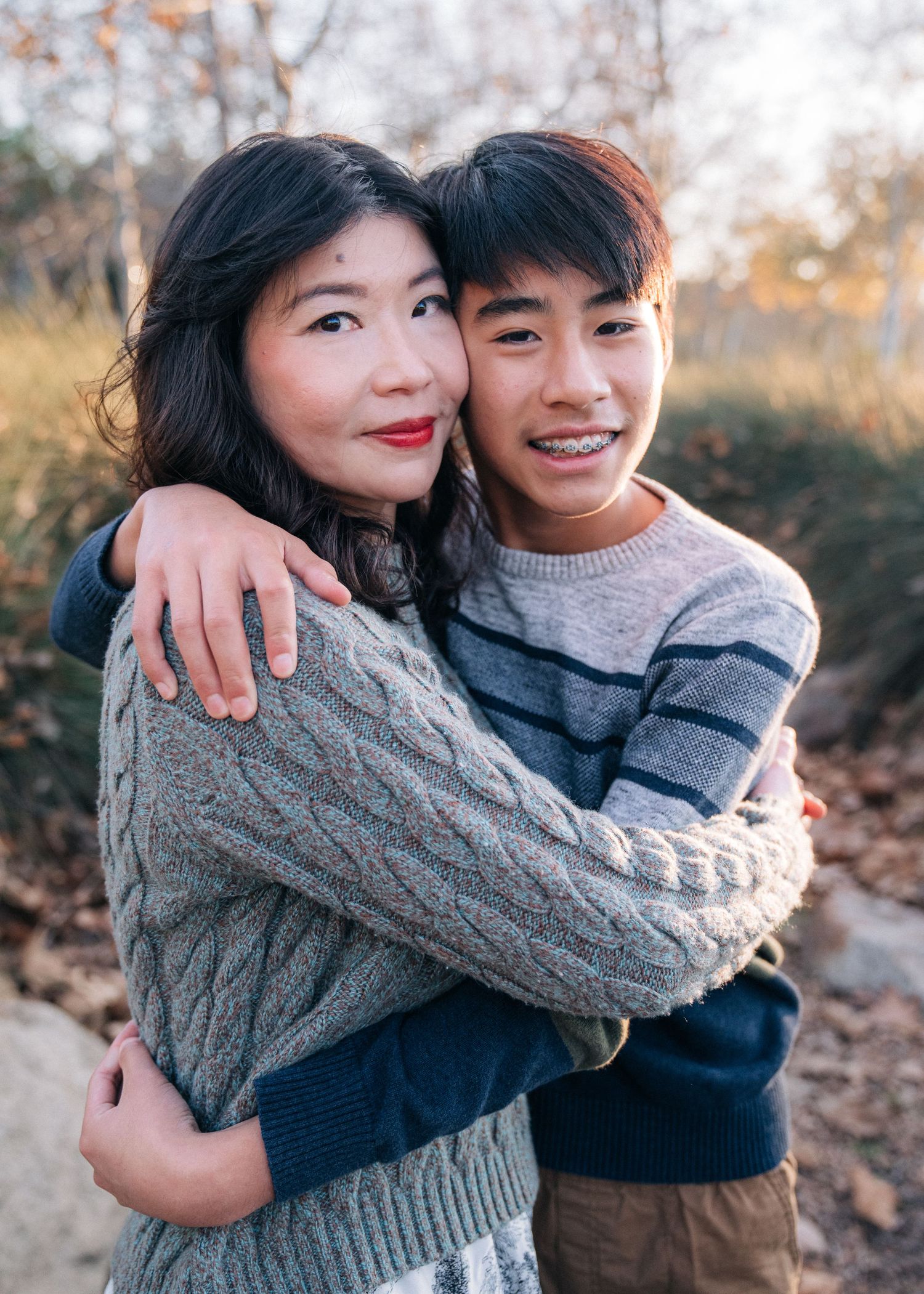 Young couple in warm sweaters embrace outdoors during autumn with golden sunlight and fallen leaves in background.