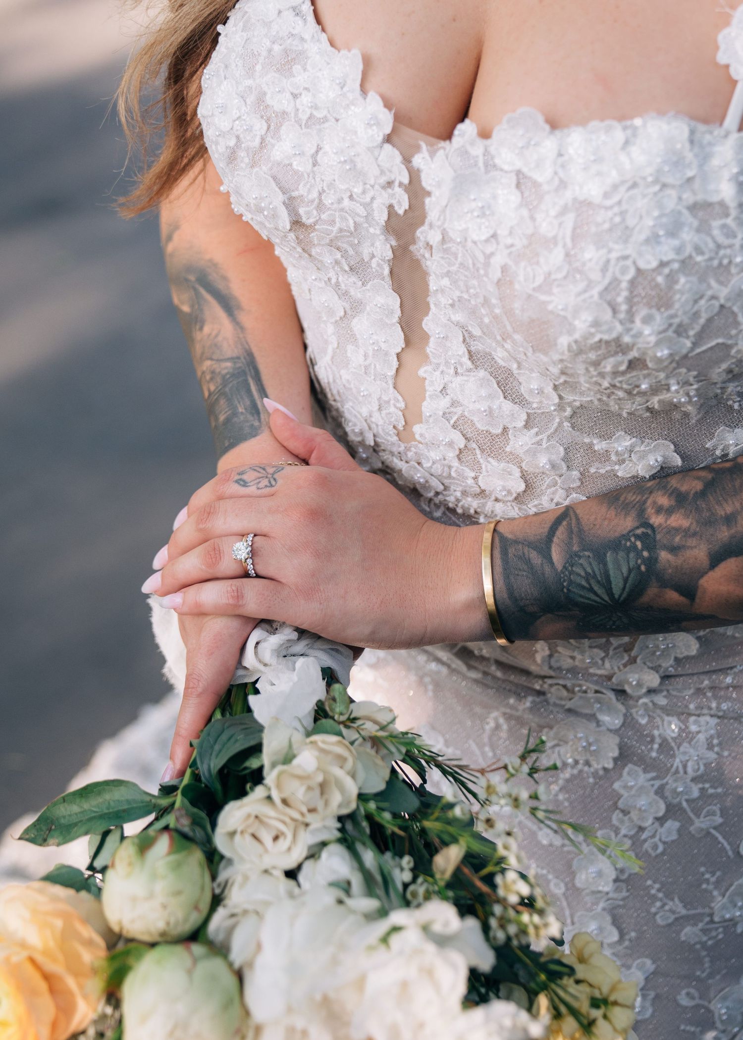 Close up detail of a white lace wedding gown bodice with tattooed arms holding a soft pastel bouquet.