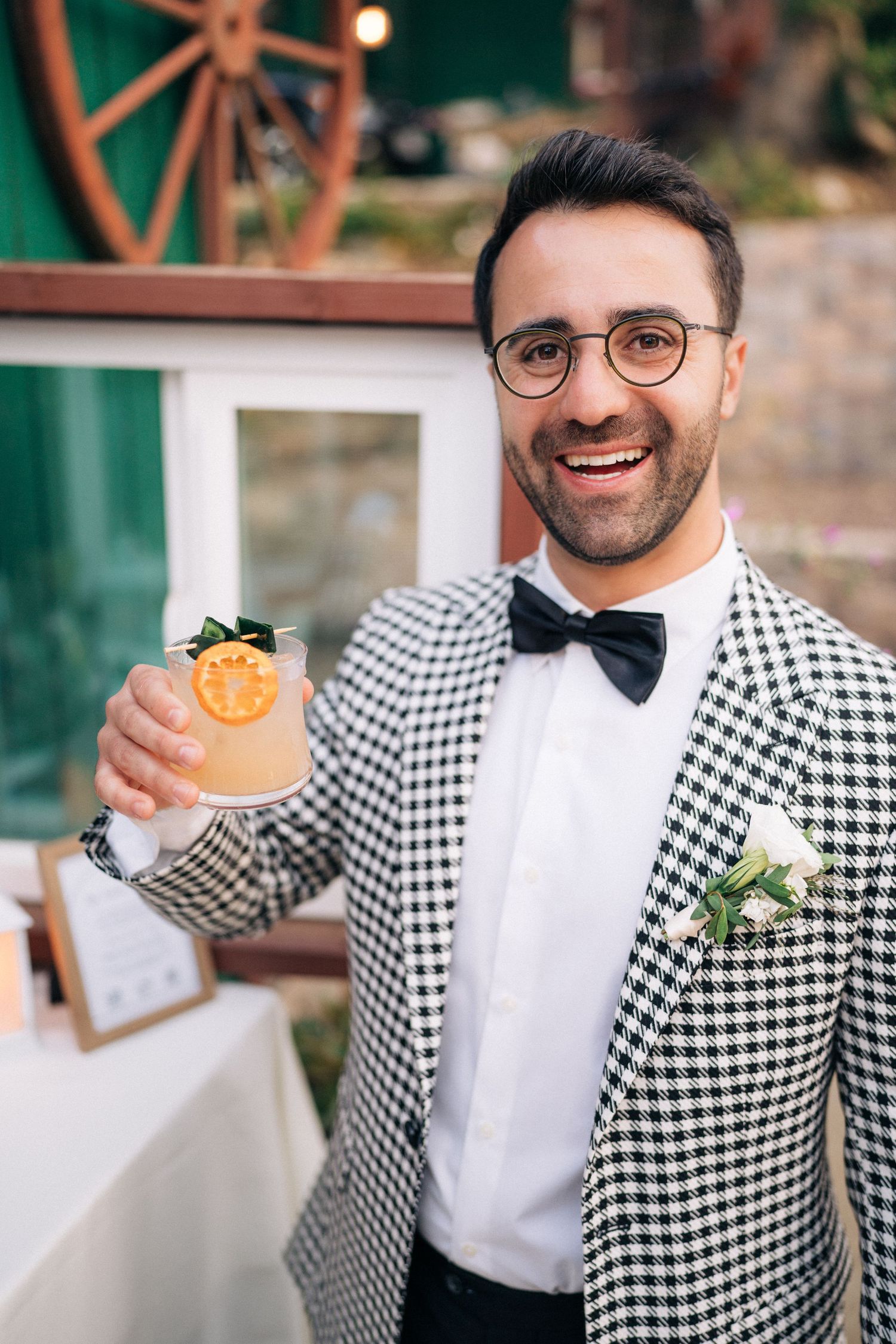 A groom in a checkered suit and bow tie holds up a cocktail while smiling at an outdoor reception.