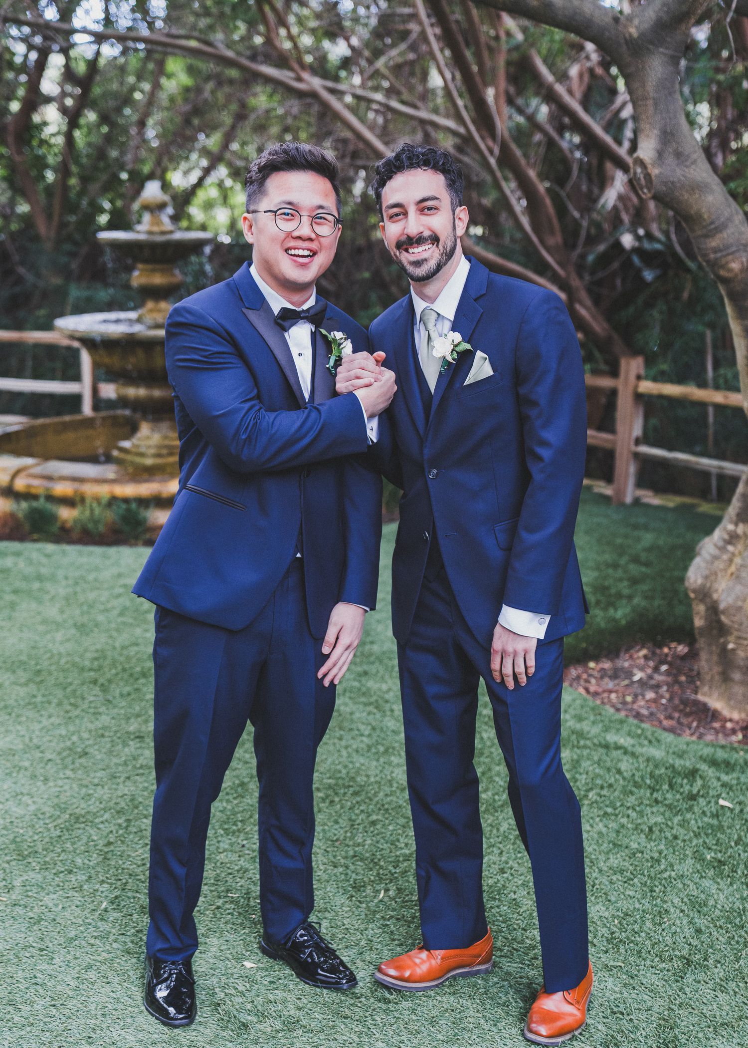 Two people in matching navy blue suits smile and pose together on grass near a garden fountain.