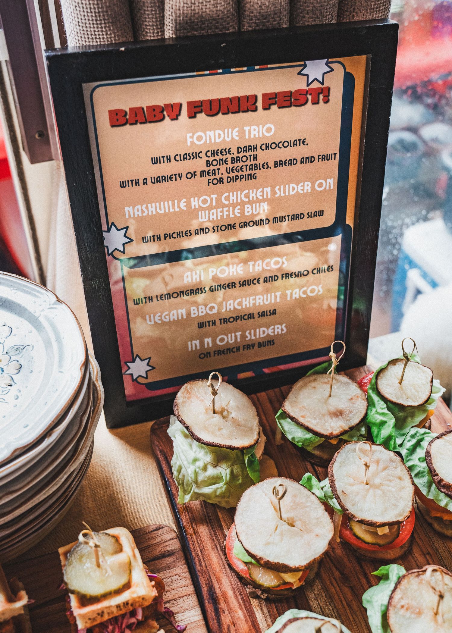 Small sandwiches on a wooden board next to a framed menu board showing food items for a pork-themed event.