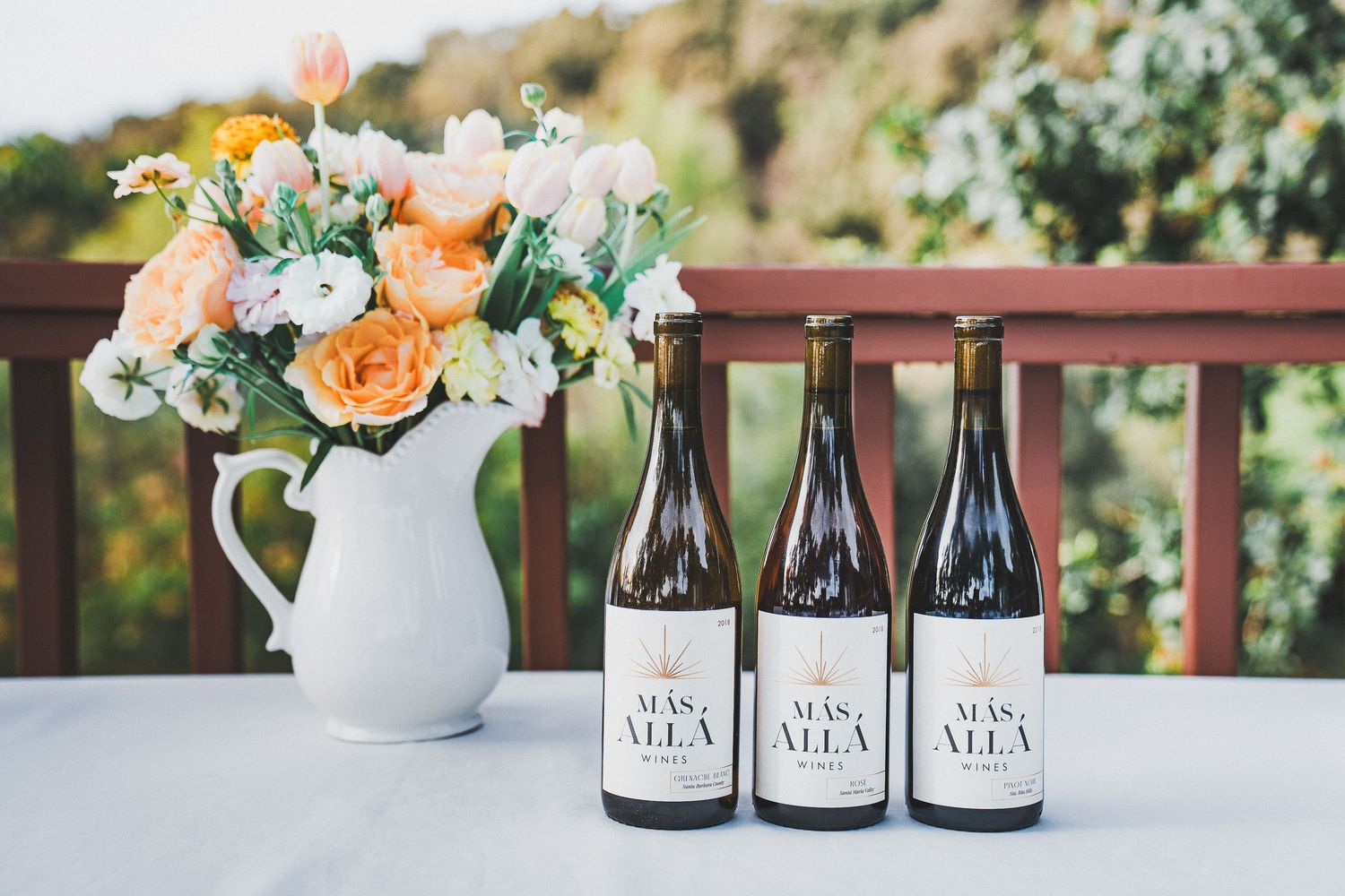 A white pitcher filled with peach and white flowers sits beside three wine bottles on an outdoor deck railing.