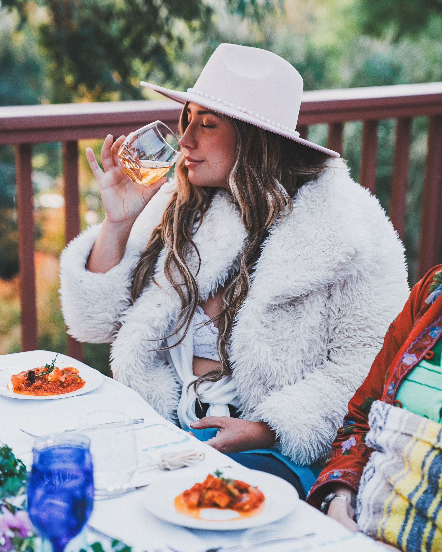 Someone in a white fuzzy coat and wide-brim hat enjoys wine while dining outdoors with plates of food on the table.