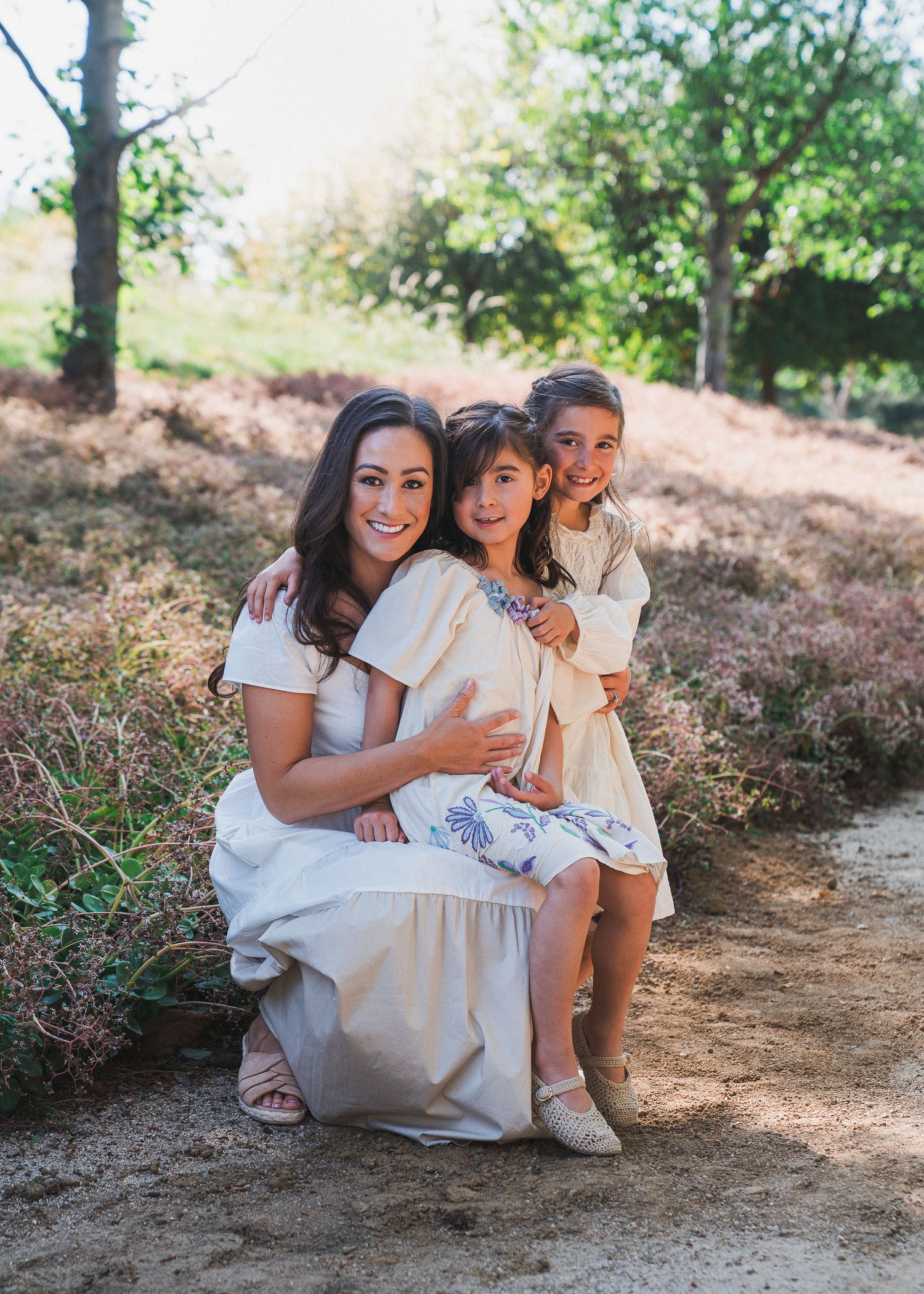 Three people pose together on a dirt trail surrounded by blooming purple wildflowers and trees on a sunny day.
