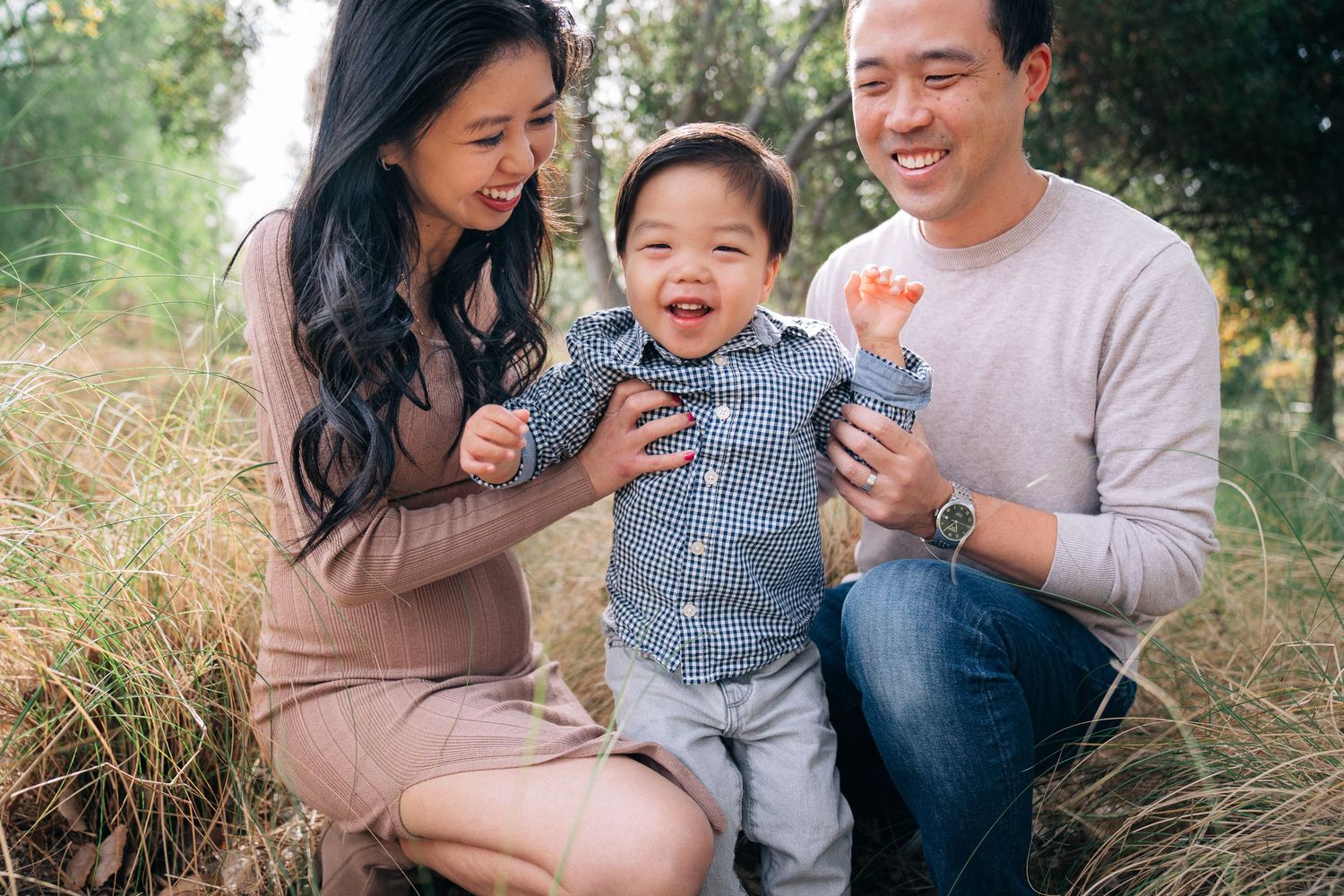 A family of three shares a joyful moment together while sitting in tall grass on a sunny day.