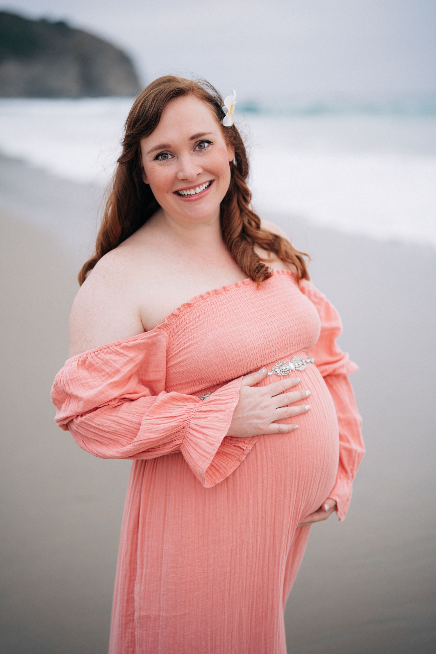 A woman in a coral dress poses for maternity photos against a misty beach backdrop.