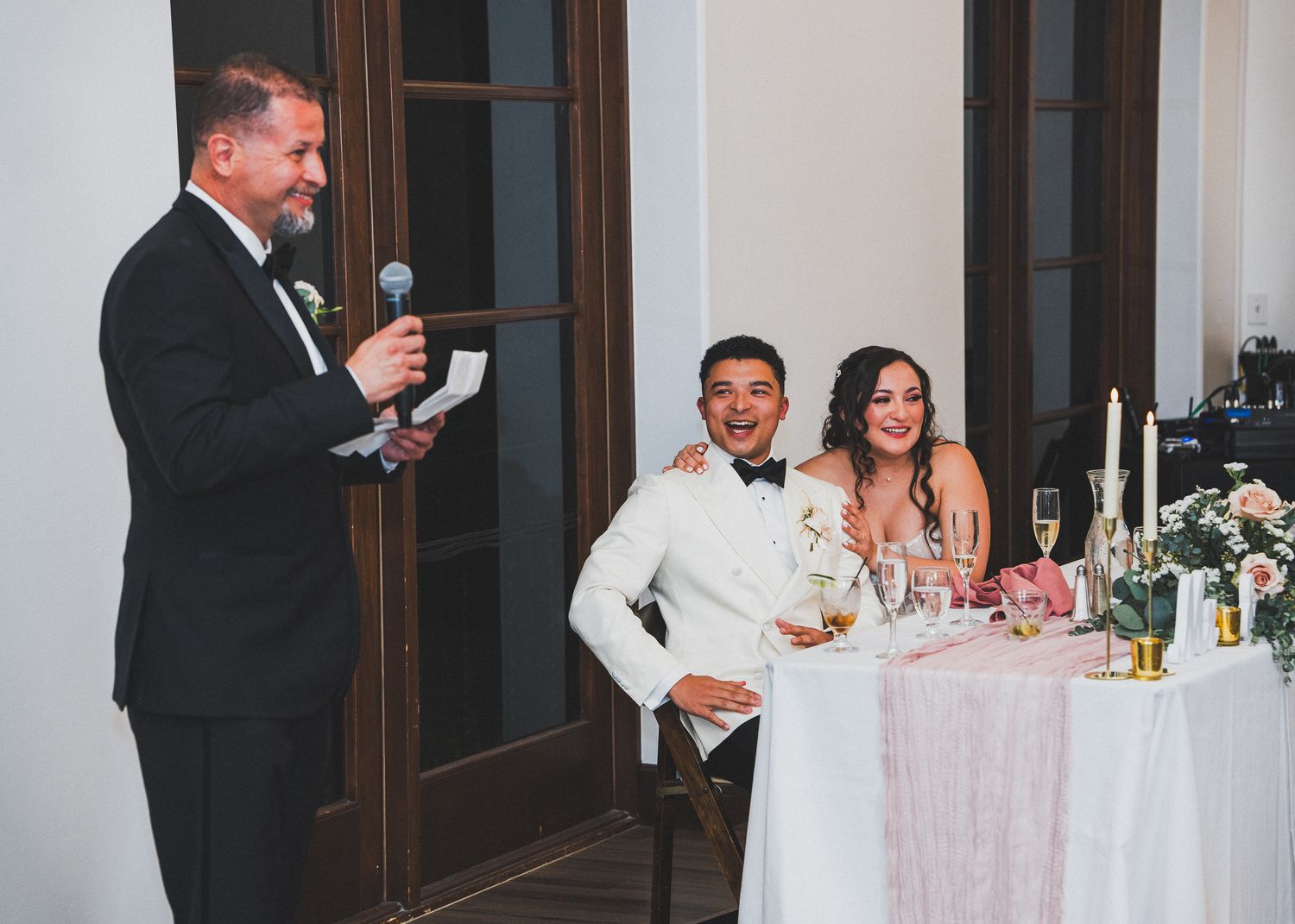 Wedding guests enjoy speeches and toasts at an elegant reception with candles and floral decor at the head table.