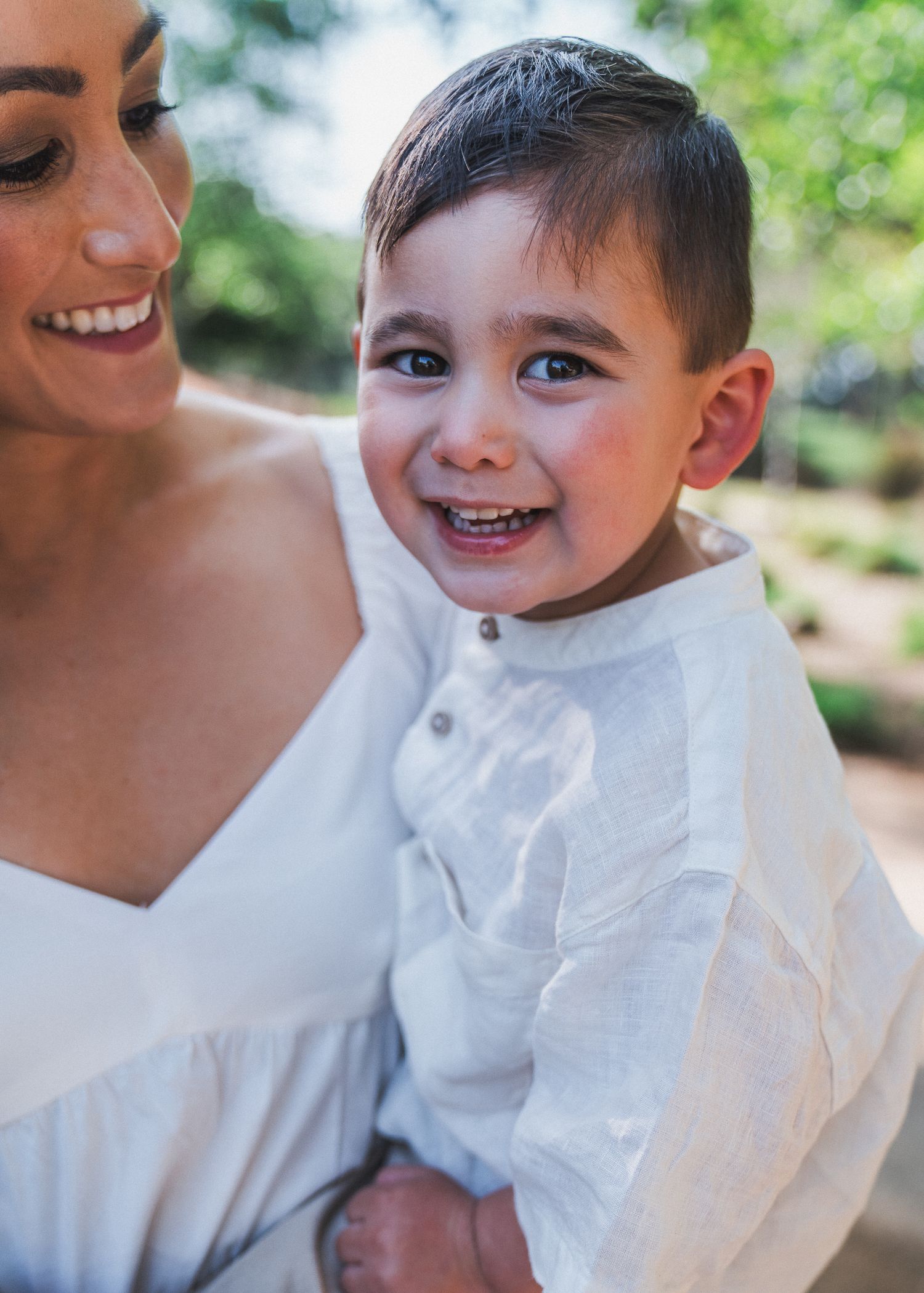 A child in a white long-sleeved shirt laughs joyfully while being held in a bright outdoor setting.