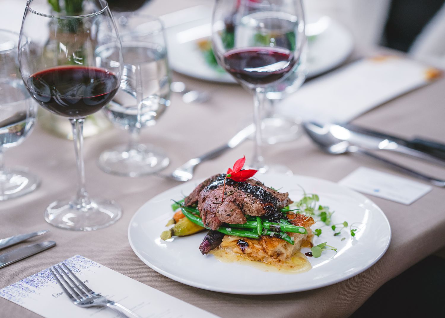 Elegant plated meat dish with vegetables and wine glasses on a formal dining table setting.