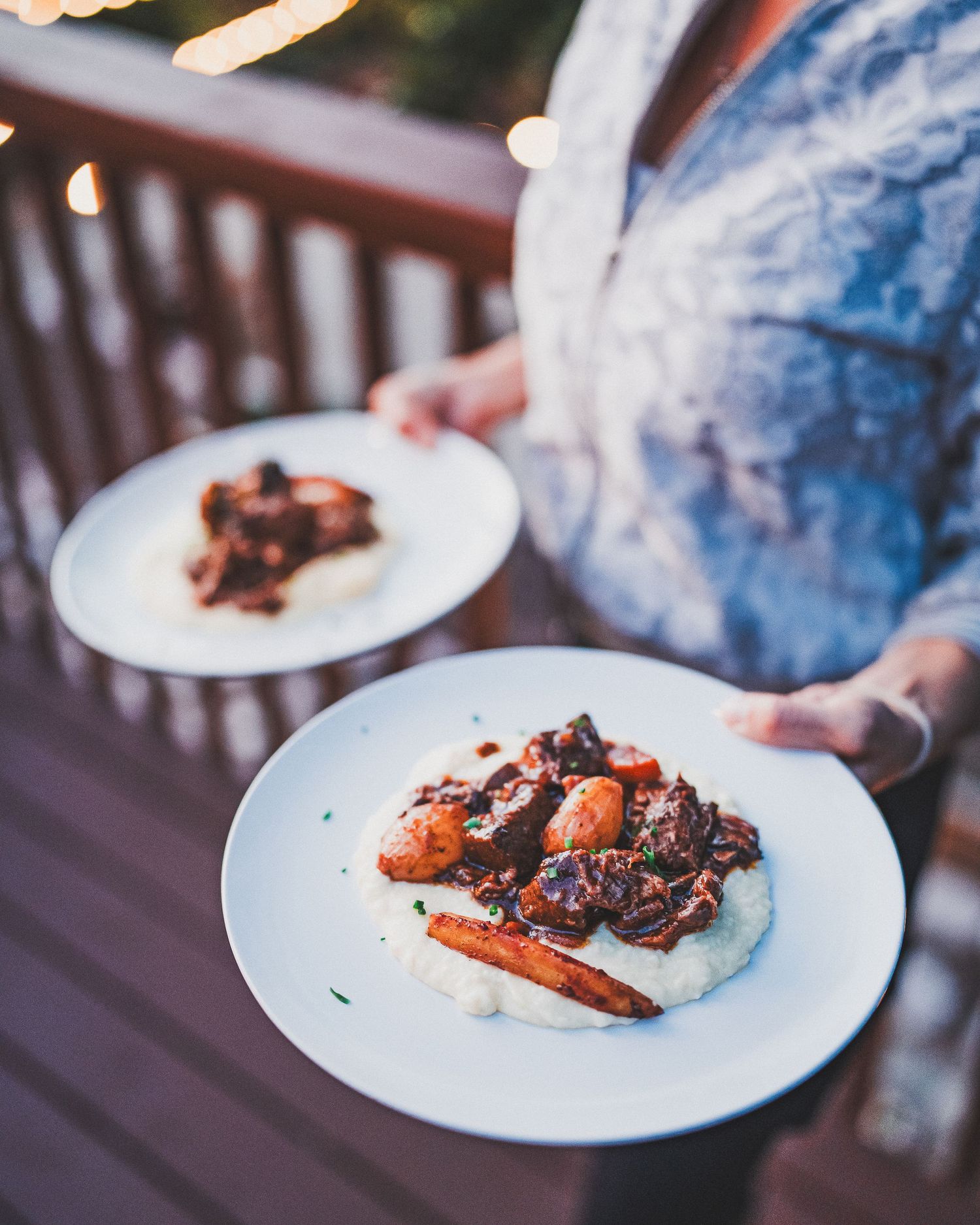 Two plates of braised meat and vegetables served on white dishes against a wooden deck railing.