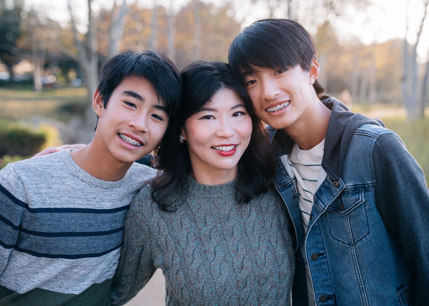 Three friends smile warmly together outdoors on an autumn day wearing casual sweaters and denim.