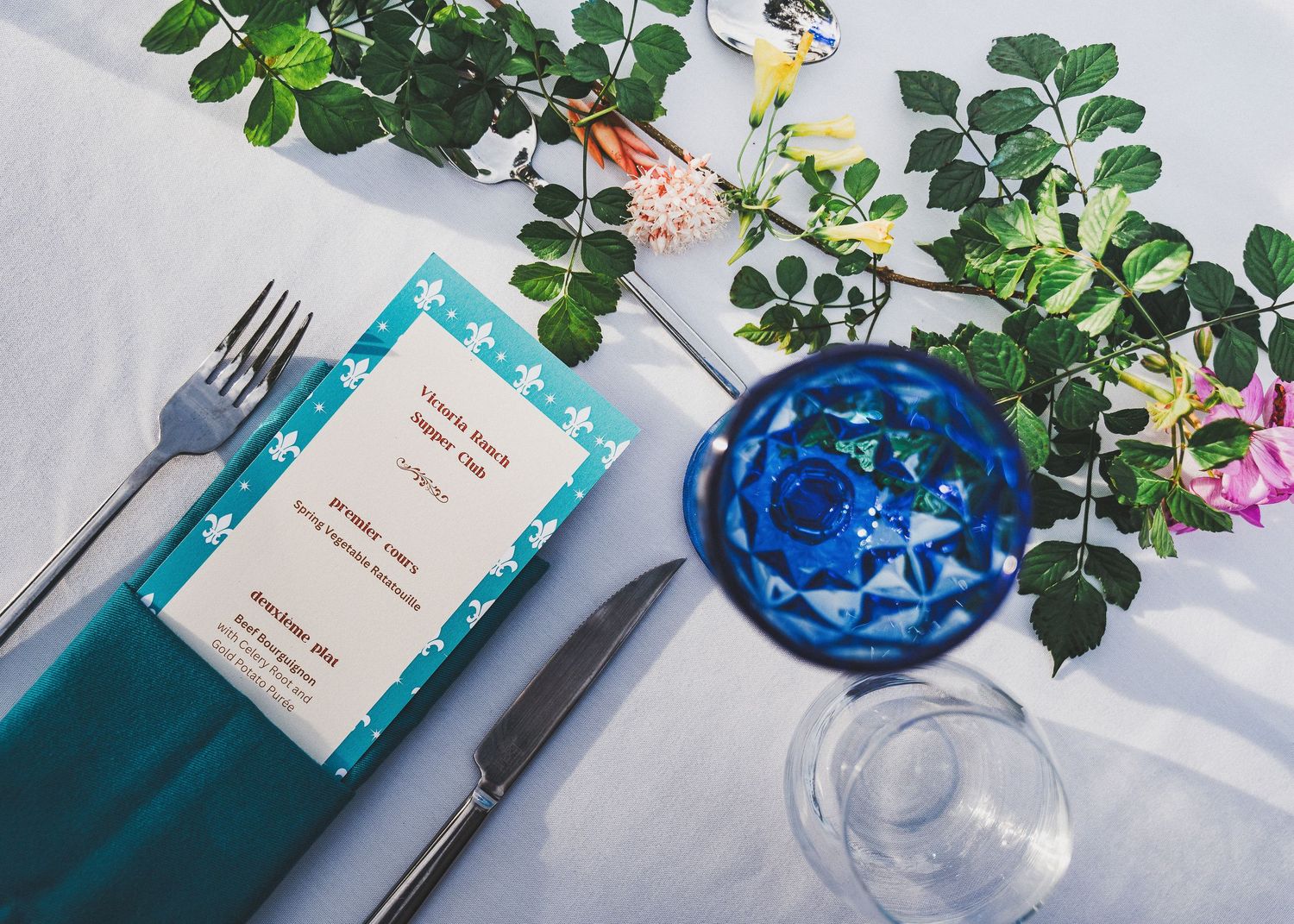 Place setting with turquoise napkin, menu card, silverware and blue glass vase containing delicate floral arrangement.