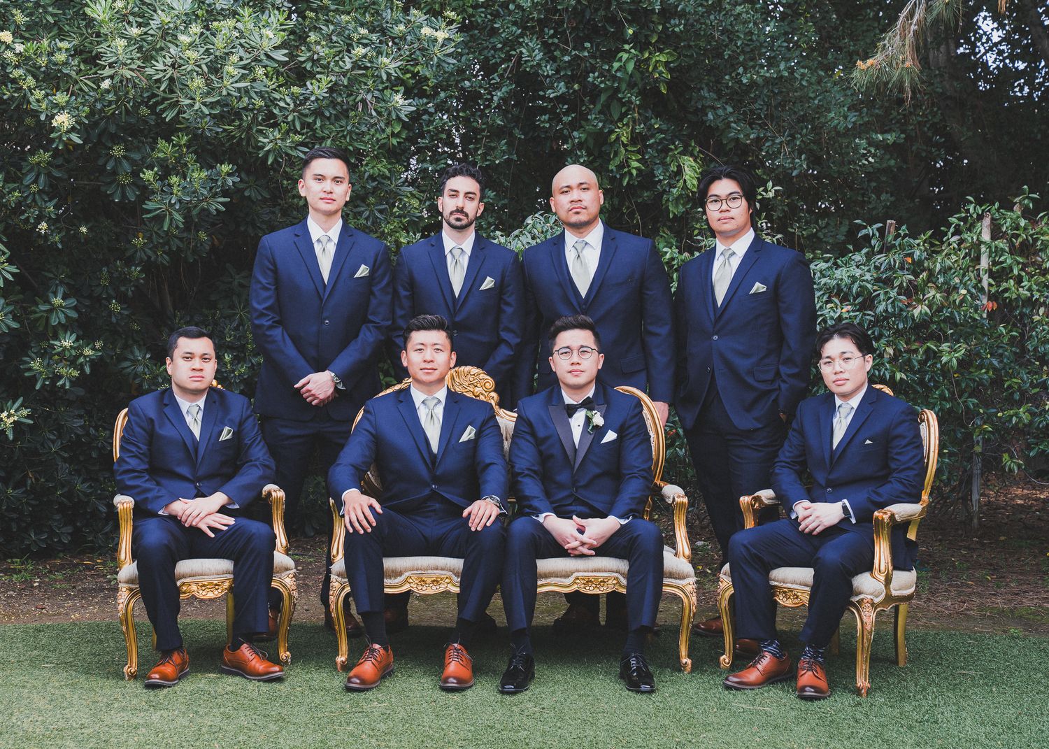 Wedding party in navy blue suits sit casually on wooden chairs outdoors against a green garden backdrop.