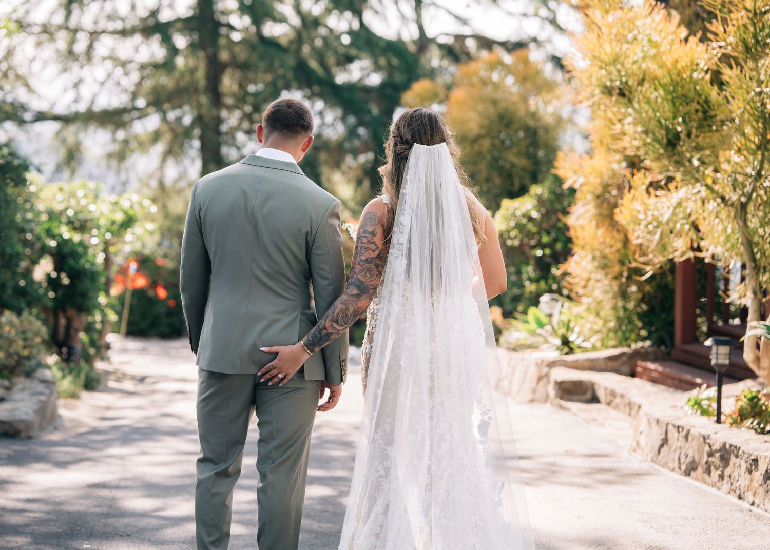 A couple in wedding attire walk along a sunlit garden path with golden trees and foliage in the background.