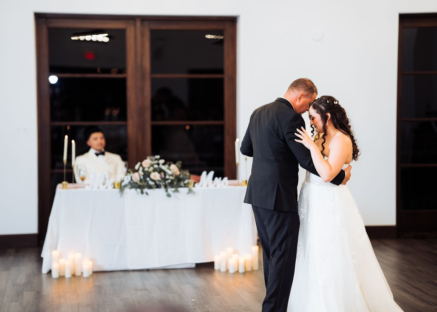 A bride and groom share an intimate moment during their first dance in an elegant reception hall with candlelit tables.