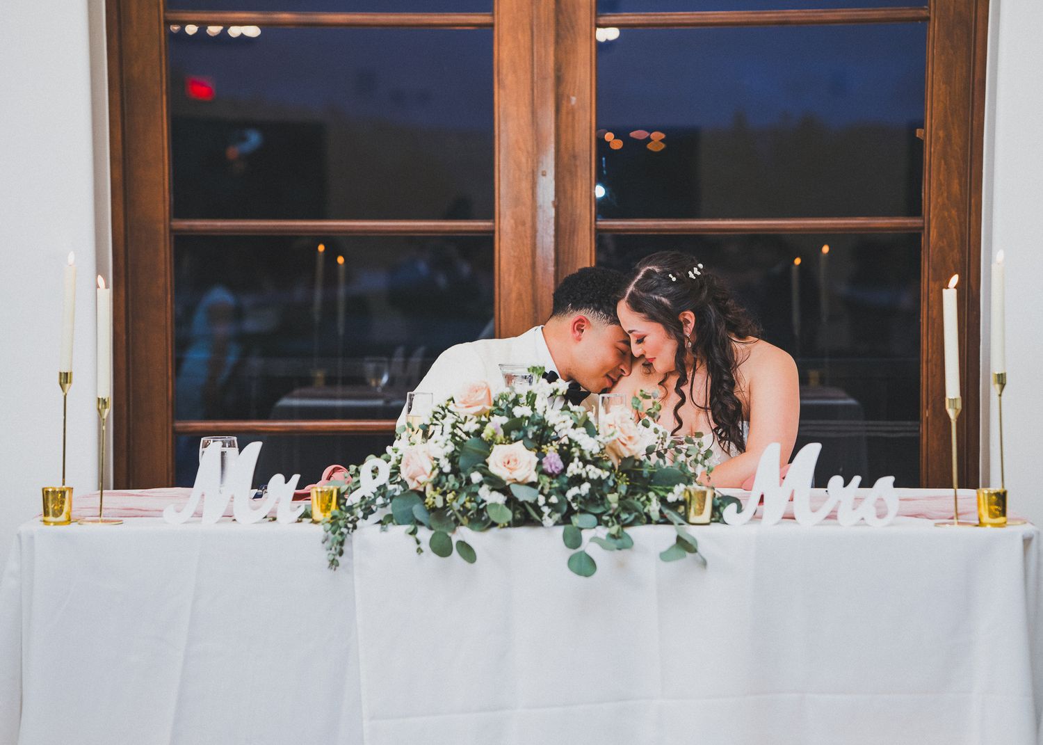 A wedding reception table setup with white linens, glassware and floral centerpieces near large windows.