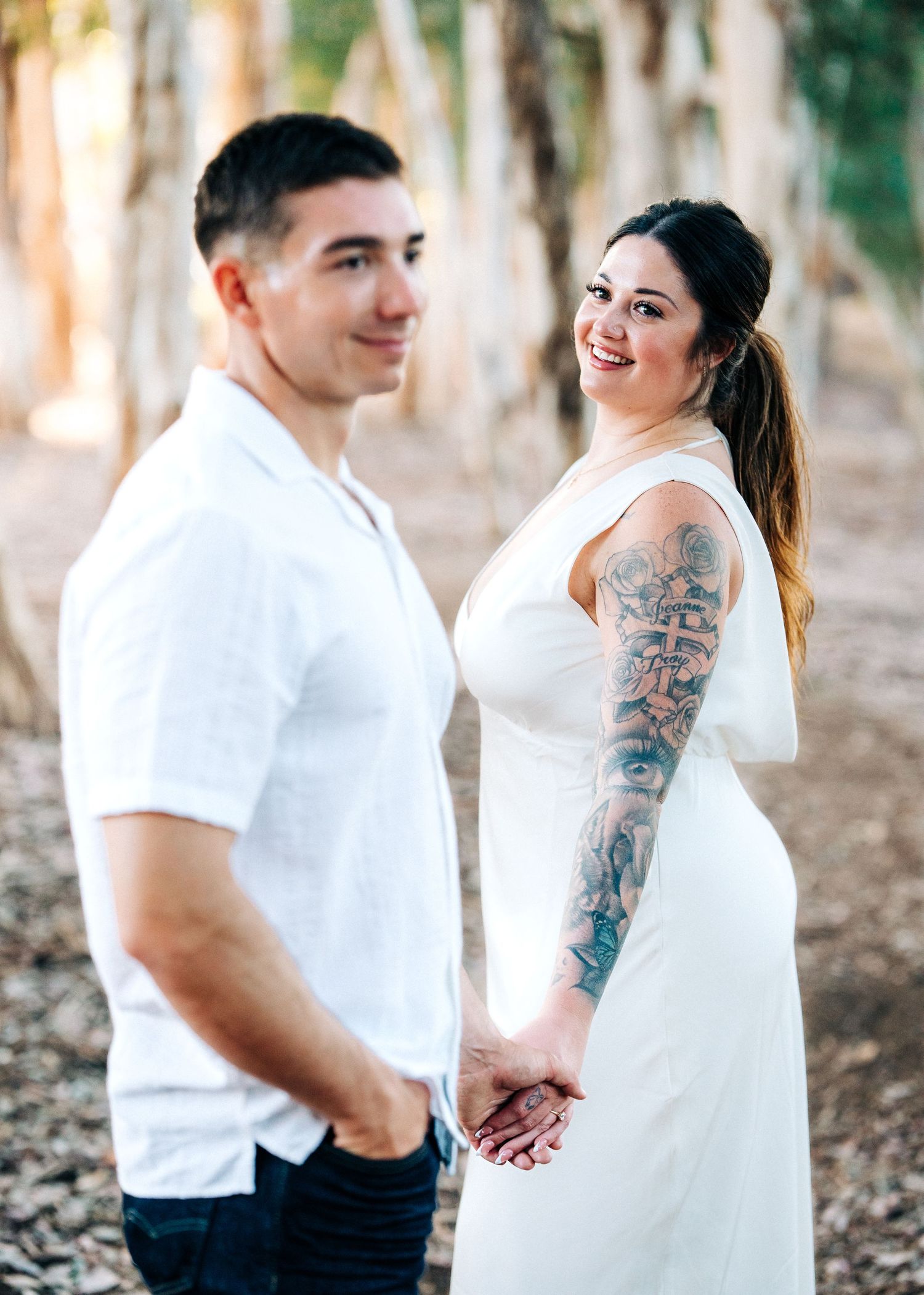 A joyful couple in white outfits holds hands while posing together in a wooded setting during a photo session.