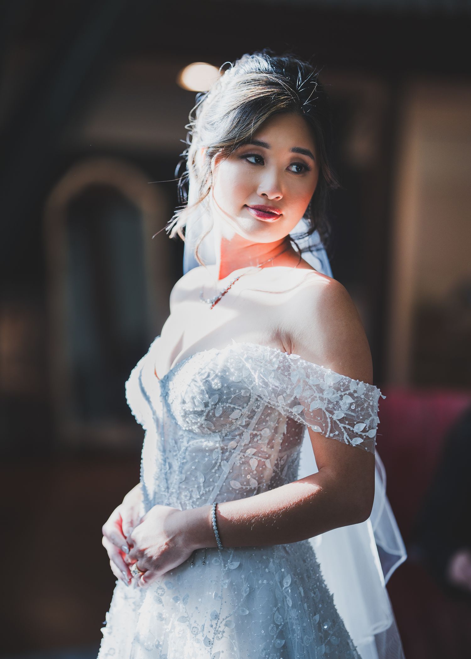 Bride in off-shoulder lace wedding gown poses in dramatic lighting.