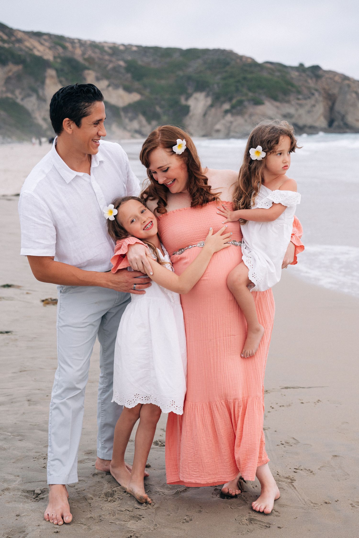 A family embraces while wearing coordinated white and coral outfits during a beach photoshoot at sunset.