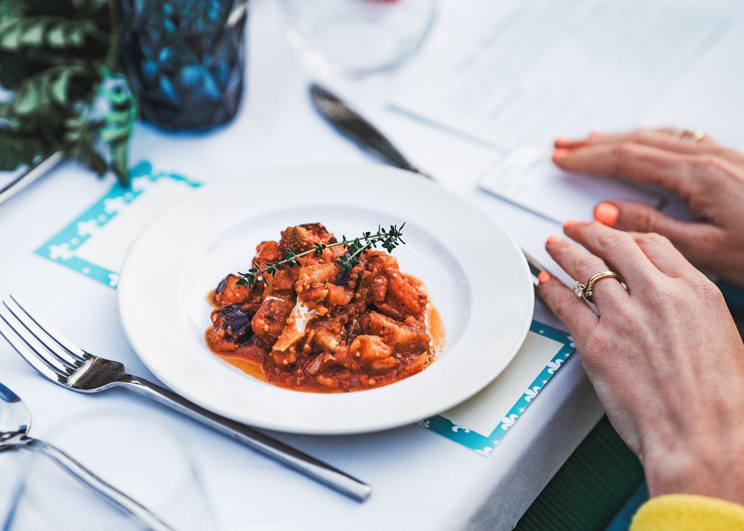 A plate of pasta in rich tomato sauce garnished with fresh herbs sits on a white table beside hands holding cutlery.