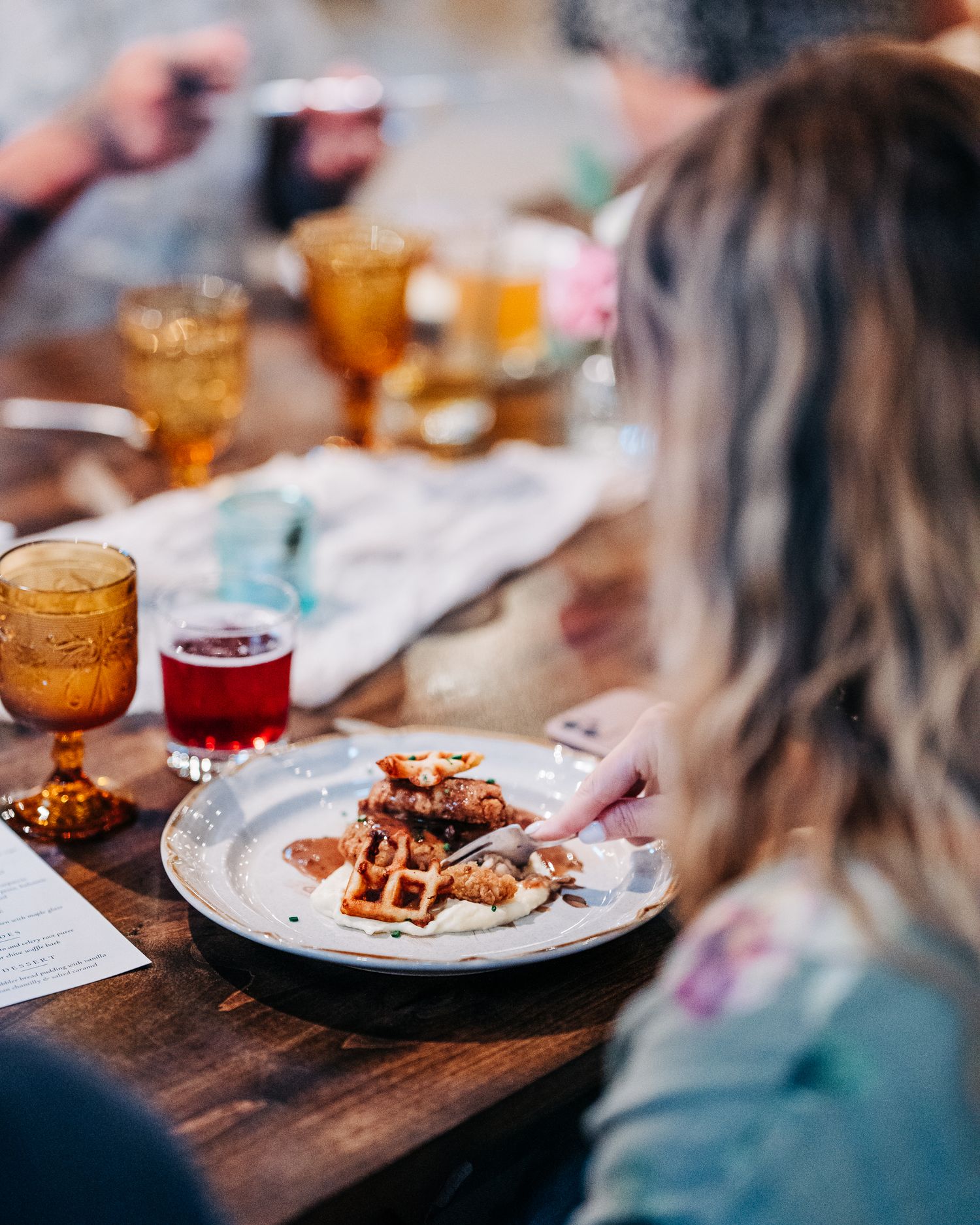 A plate of waffles with toppings is served on a wooden dining table with colorful drinks in amber glassware.