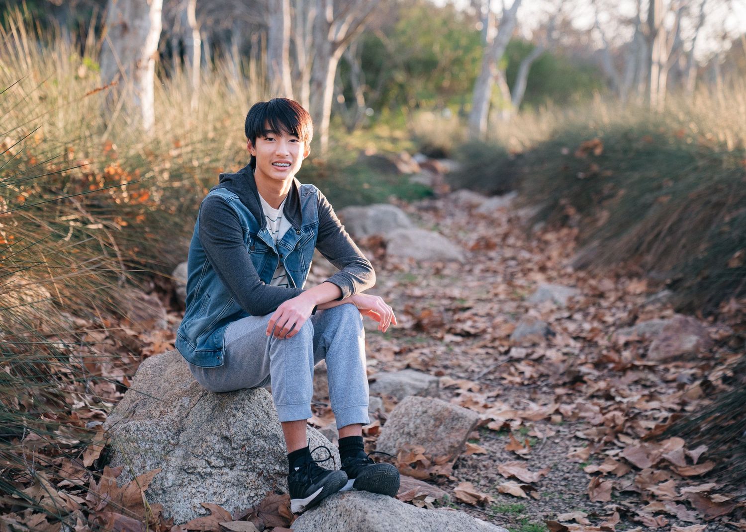 A person in denim sits casually on a large rock along a leaf-covered hiking trail.