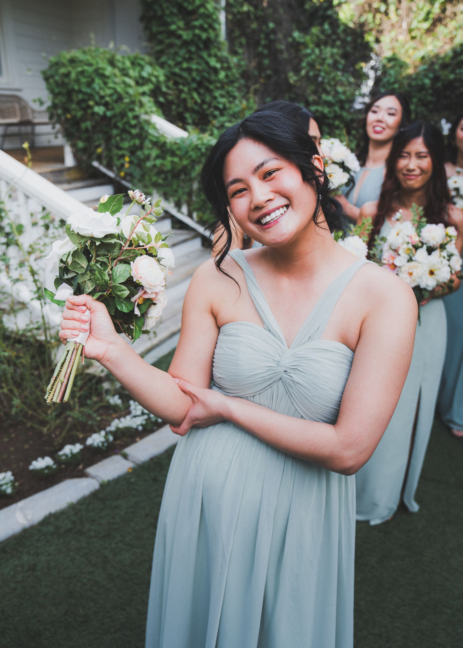 A bridesmaid in a flowing pale blue gown holds a white flower bouquet while smiling joyfully at an outdoor wedding.