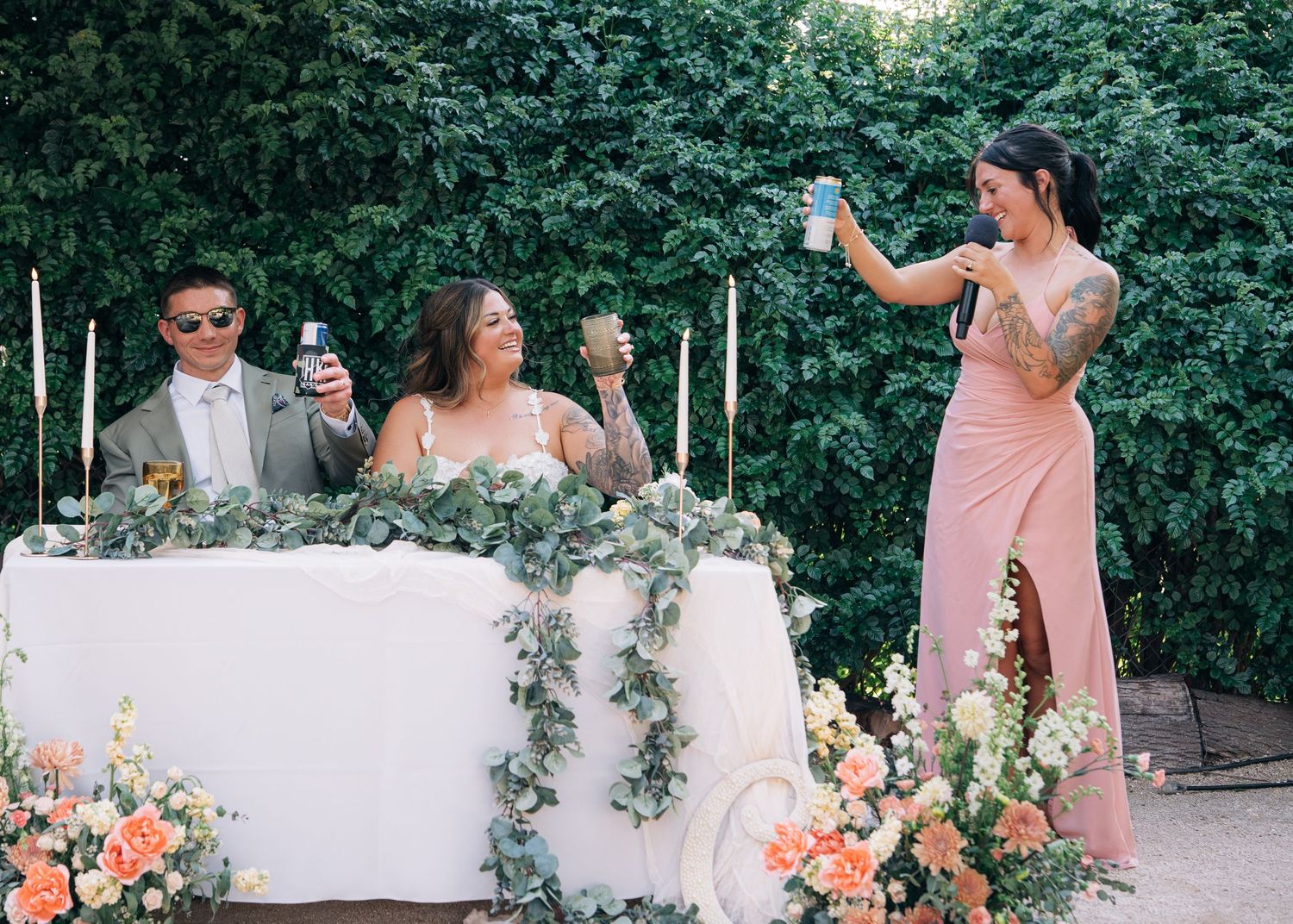 Wedding reception guests seated at a floral-decorated white table with a person in pink giving a toast outdoors.
