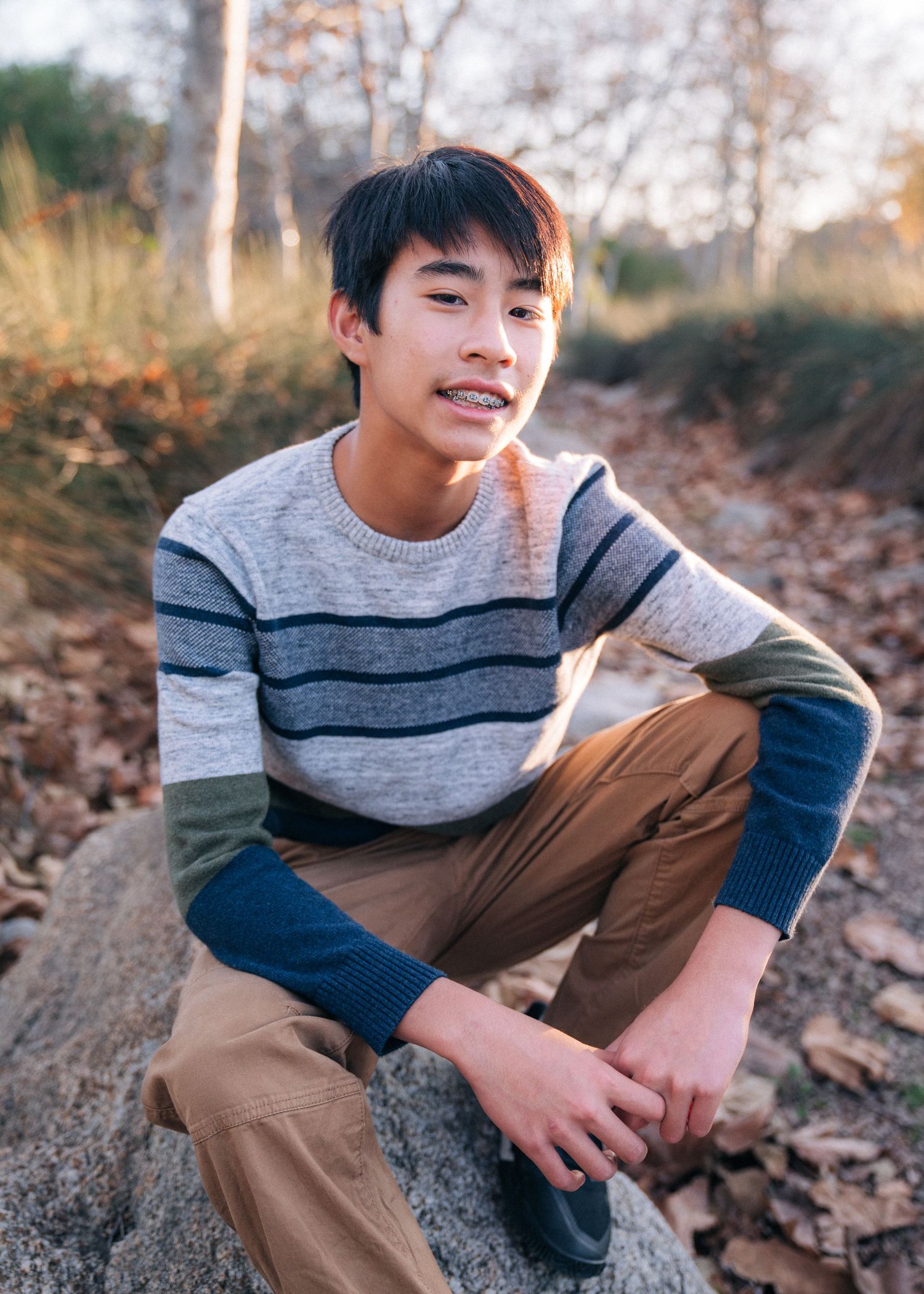 A person in a striped sweater poses on a rock amid fallen autumn leaves on a nature path.