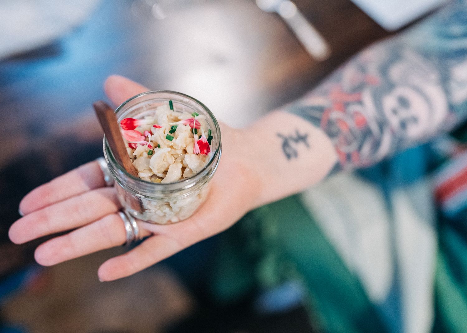 Glass jar containing creamy dessert with red garnish held by tattooed hand.