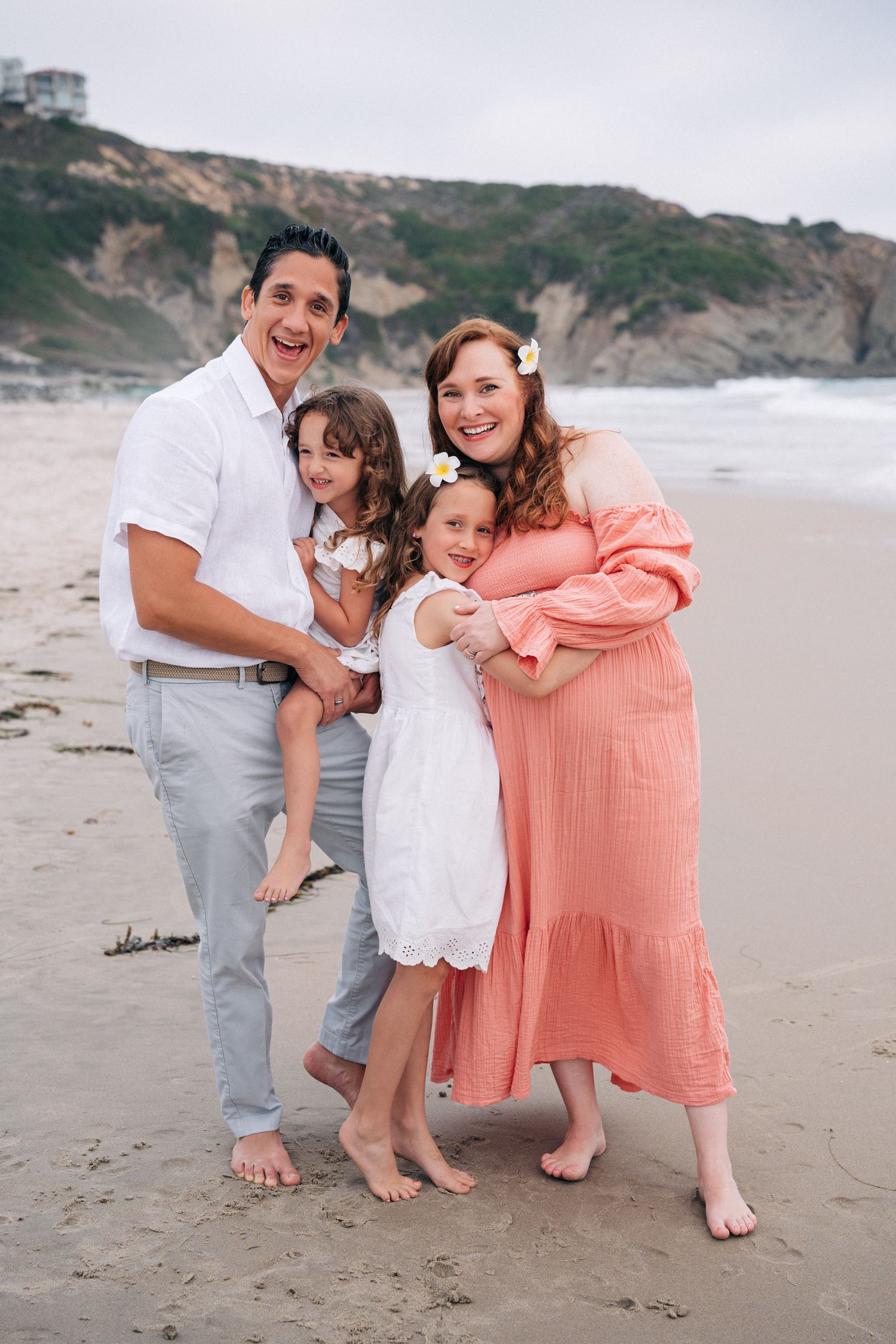 Family dressed in white and coral pink poses together barefoot on a beach with cliffs in the background.