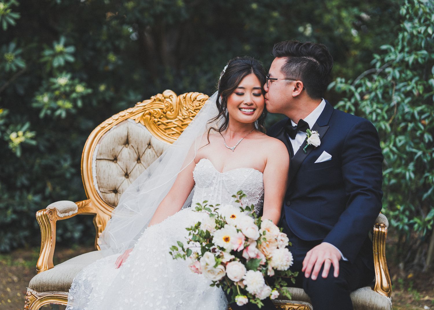 Newlyweds share intimate moment on vintage gold settee with floral bouquet.