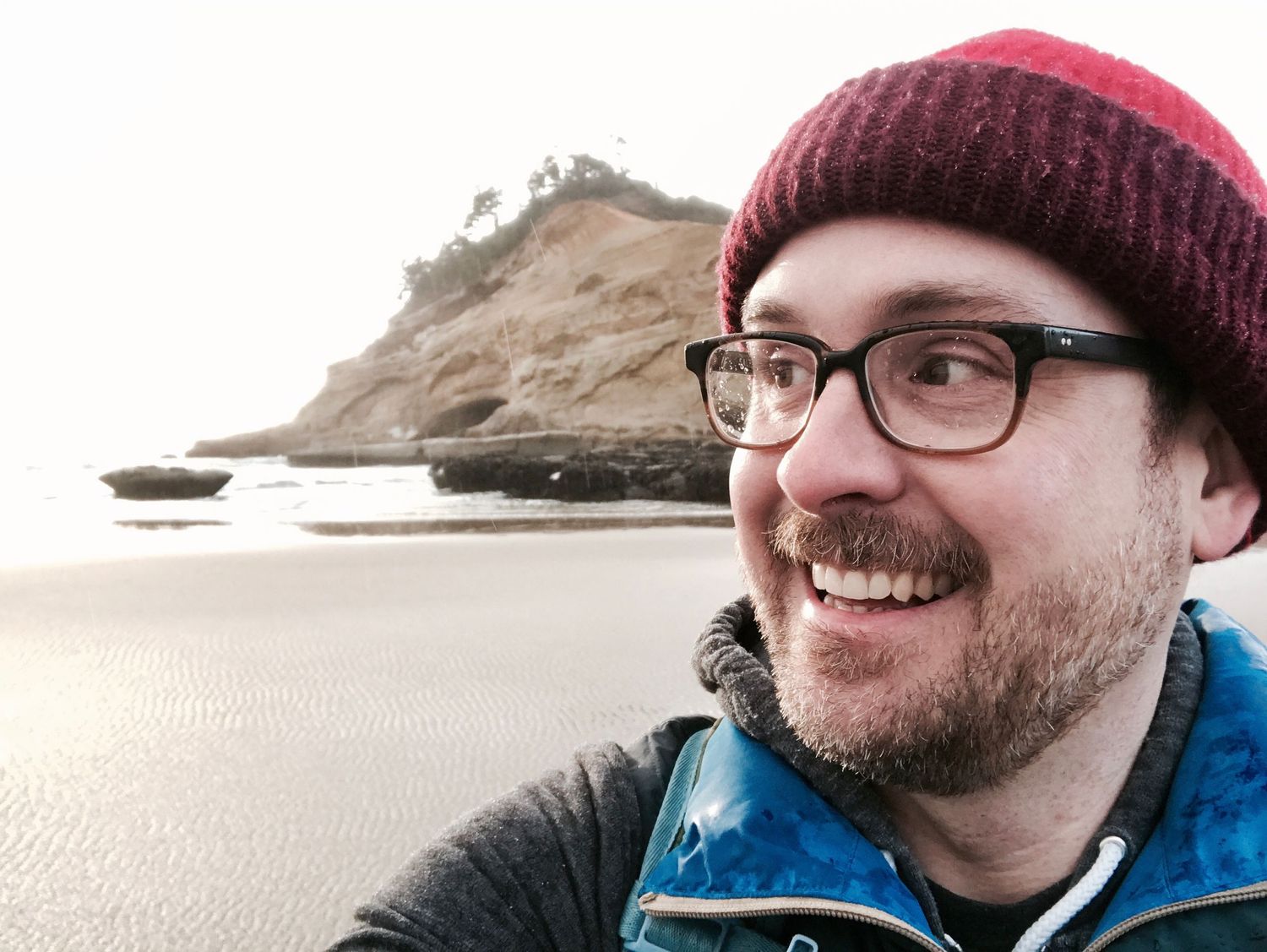 Someone wearing a burgundy beanie and glasses stands on a rocky beach during overcast weather.