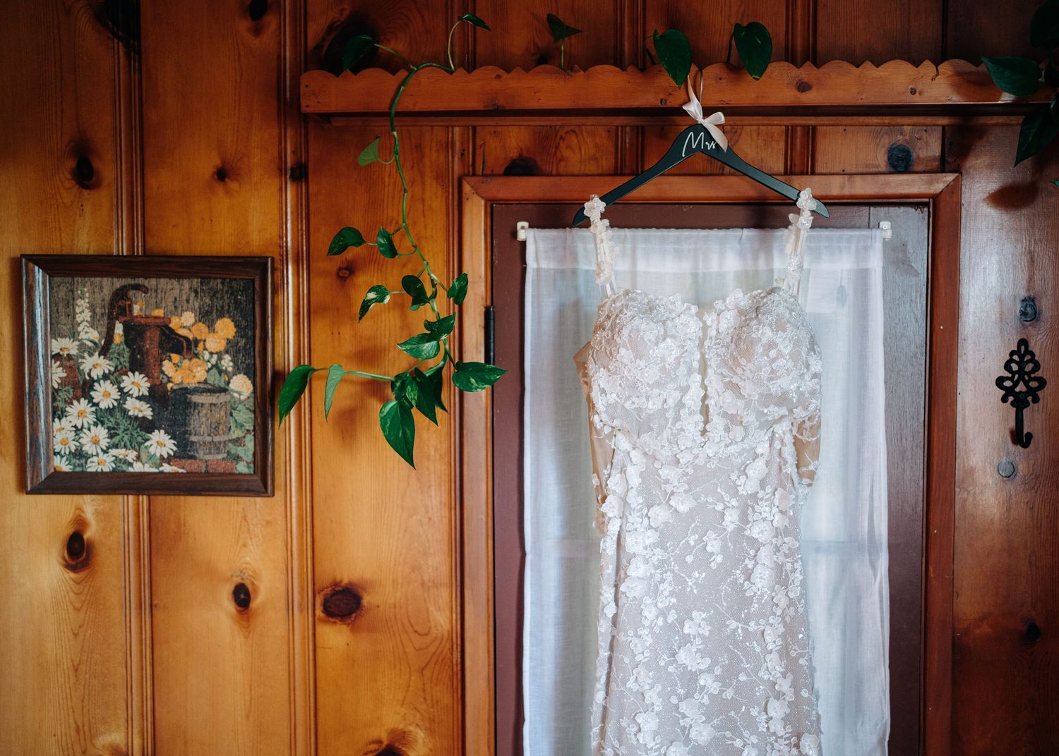 A delicate lace wedding dress hangs on display against rustic wooden paneling with decorative florals.