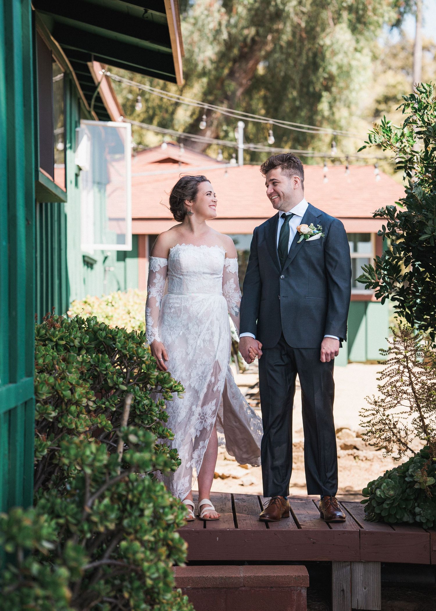 A romantic wedding photo of a couple standing on wooden decking beside a green building in natural sunlight.