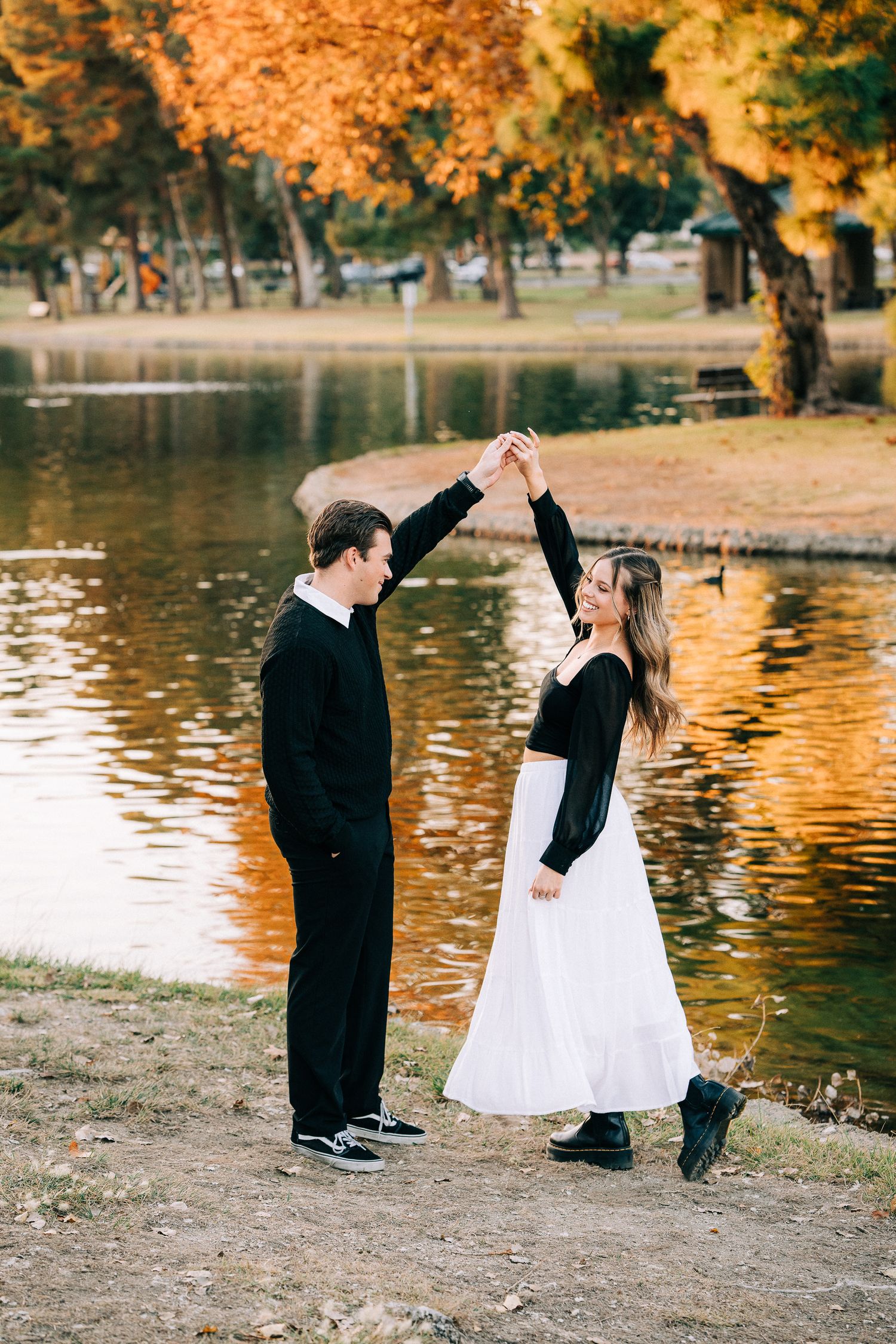A couple in black and white outfits dancing by a lake surrounded by autumn trees with orange foliage.