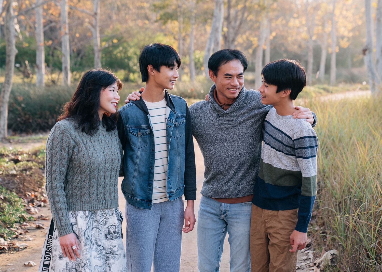 A family wearing casual sweaters and jeans smiles together while walking on a path during autumn.