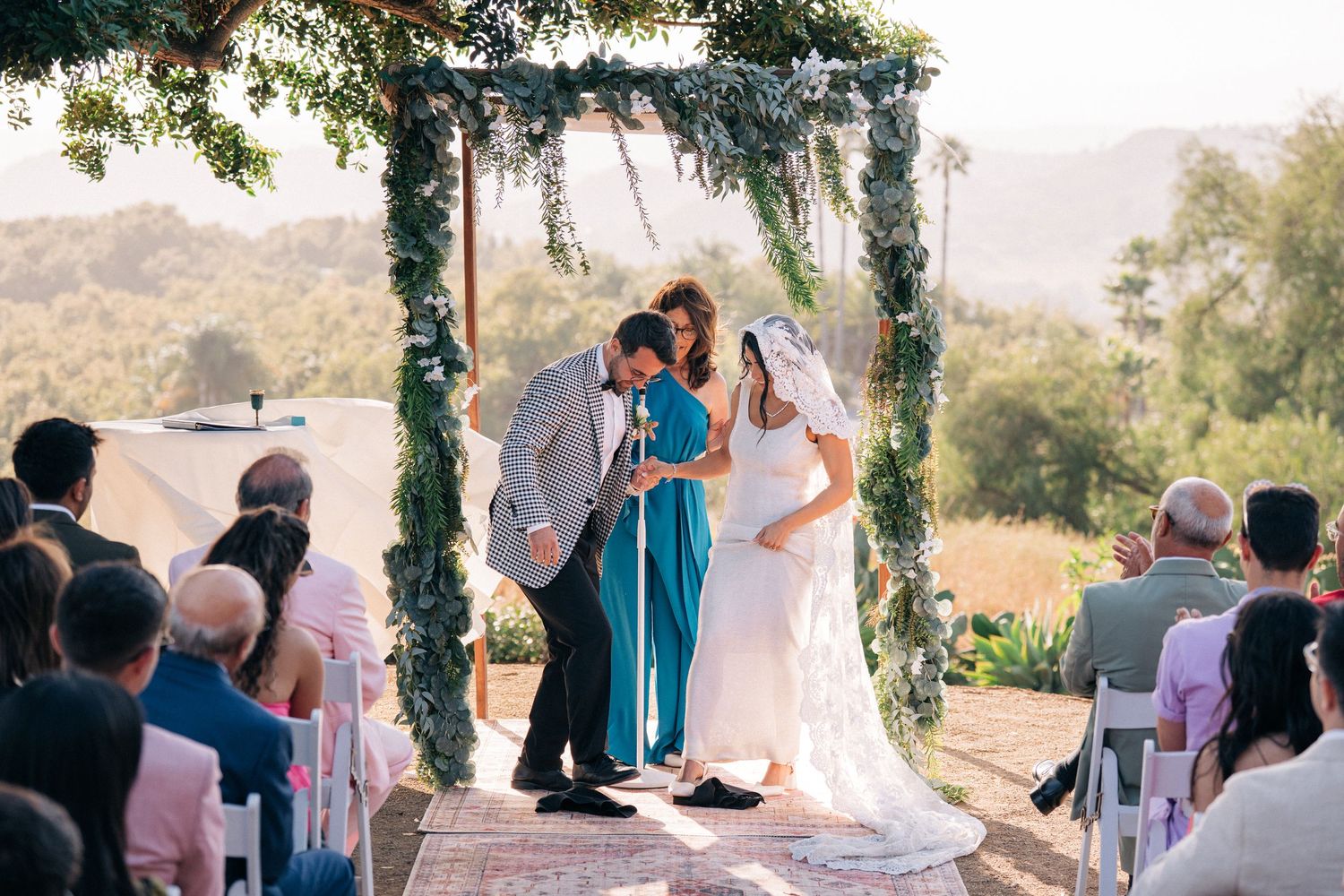 Wedding ceremony in progress under leafy arch with guests seated on white chairs.