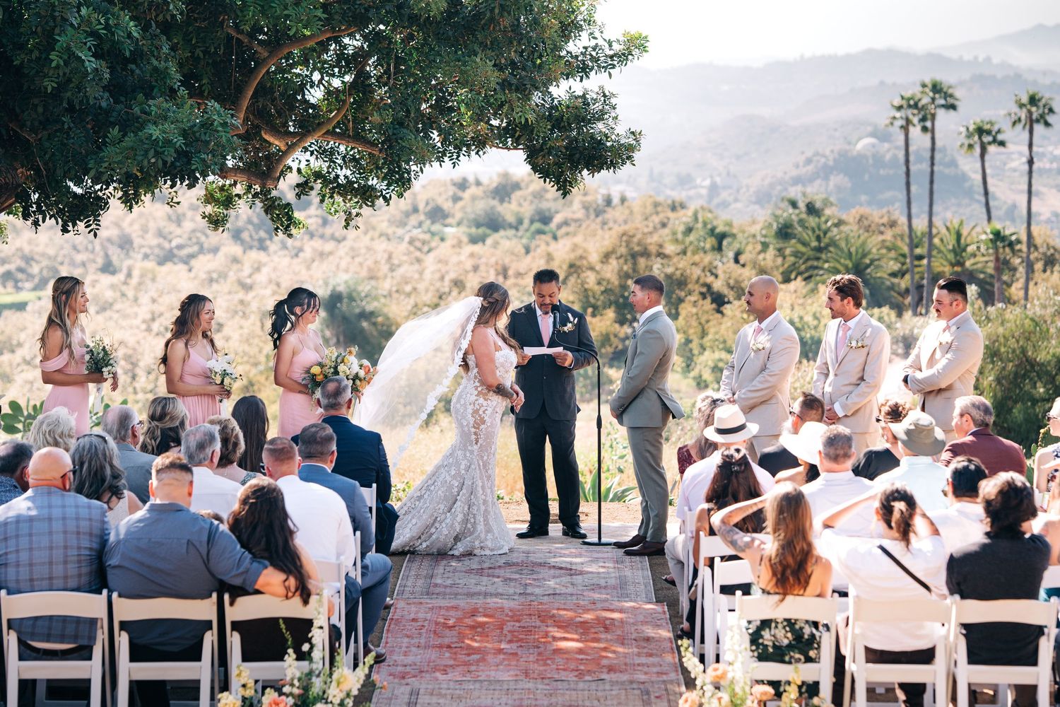 Panoramic view of an outdoor wedding ceremony with guests seated in white chairs against a desert mountain landscape.