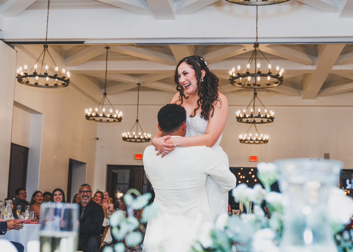 Wedding reception guests dance and socialize under elegant chandeliers in a light-filled event venue.